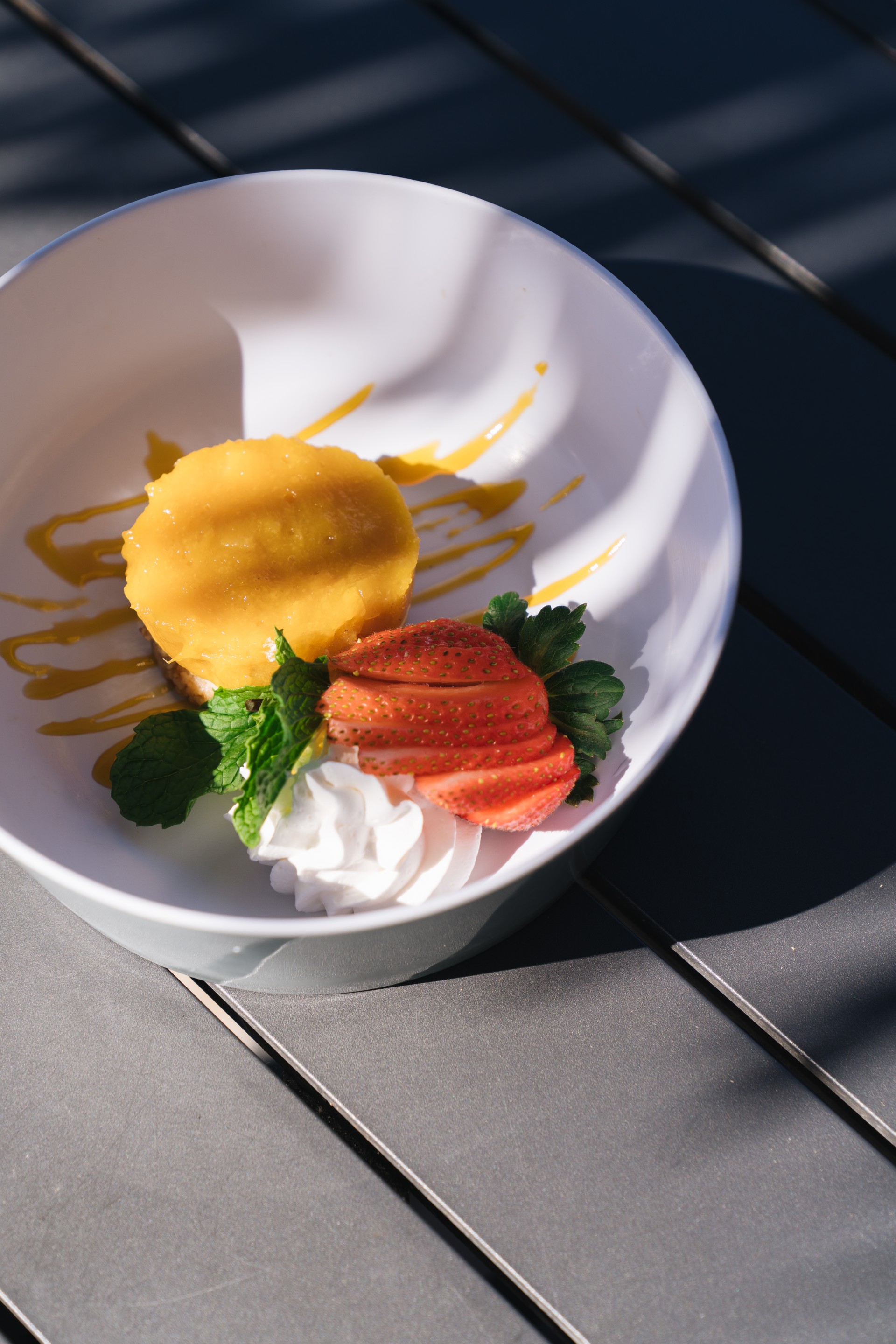 A plated tropical dessert featuring a round yellow mango tart drizzled with fruit sauce, accompanied by a fan-sliced strawberry, whipped cream, and fresh mint in a white bowl.