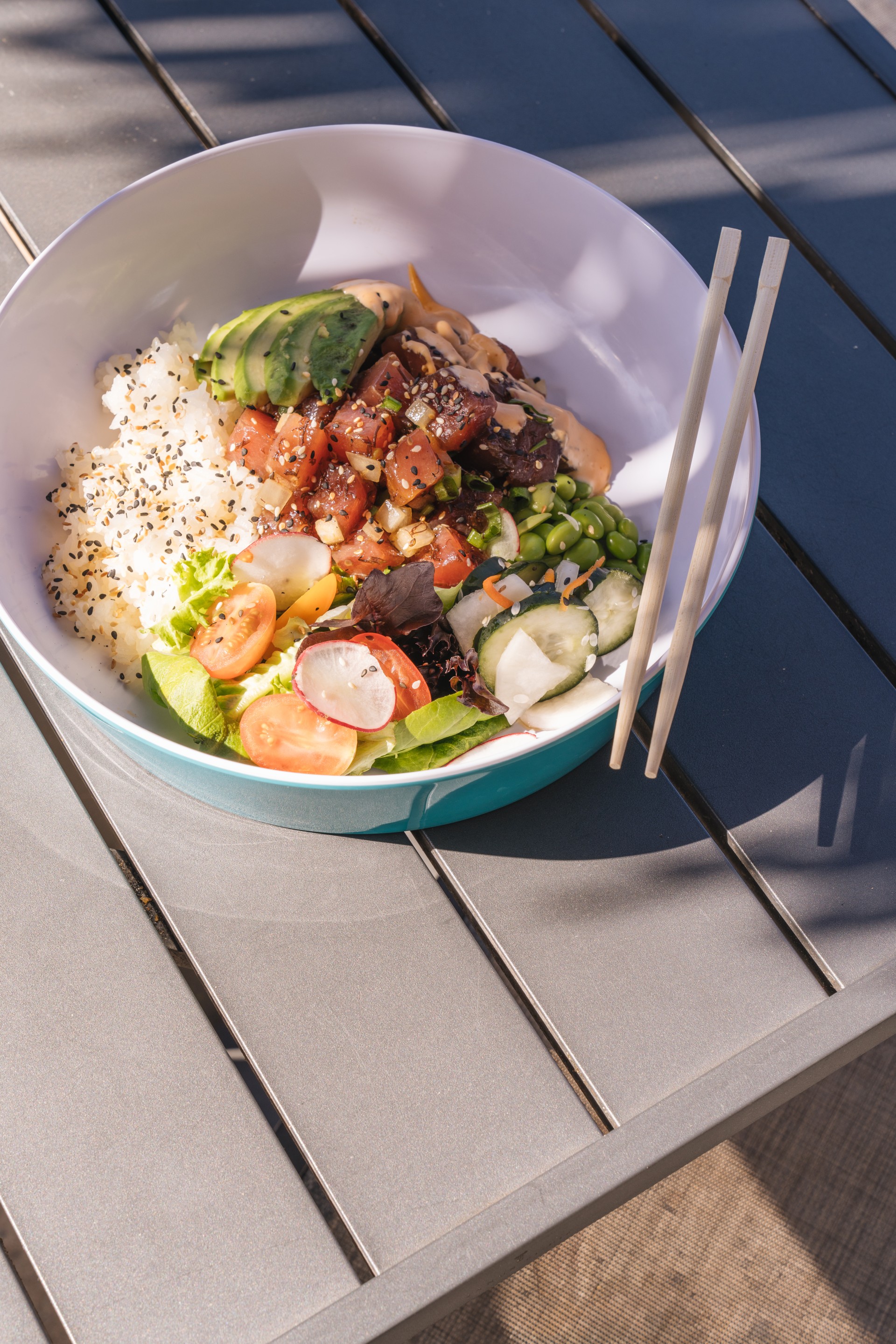 A vibrant poke bowl in a turquoise-rimmed dish featuring diced marinated tuna, sliced avocado, sesame rice, edamame, and fresh vegetables, resting on a gray slatted outdoor table with wooden chopsticks.