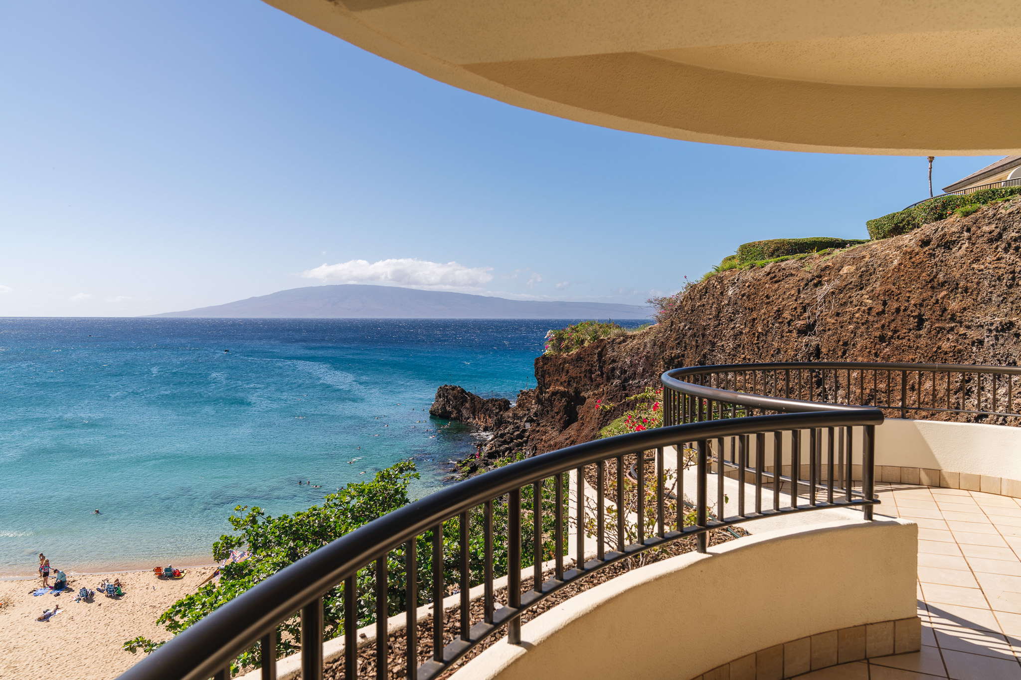 A view from a spacious curved hotel balcony with dark metal railings, overlooking a rocky coastal cliff, a sandy beach below, and vibrant blue ocean waters extending to a distant island.