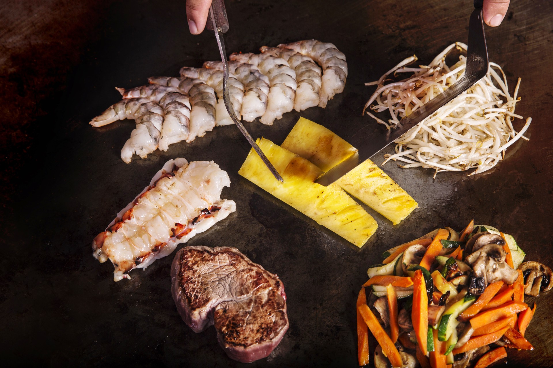 A teppanyaki chef slicing pineapple on a hot flat-top grill alongside raw shrimp, lobster tail, steak, bean sprouts, and mixed vegetables.