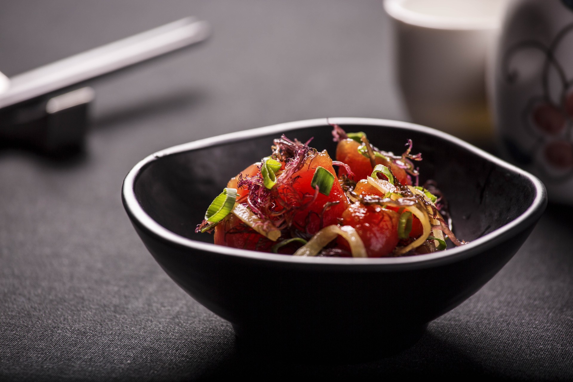 A small black bowl filled with ahi poke, garnished with shredded seaweed, sliced green onions, and sweet onions, set on a dark tablecloth with chopsticks in the background.