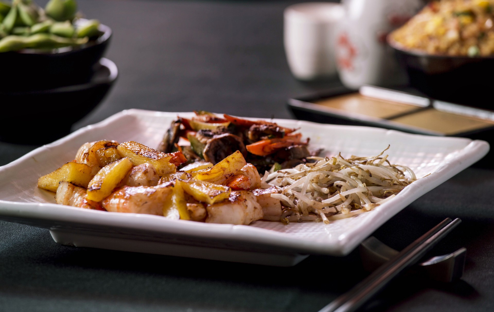 A plated teppanyaki meal on a square white ceramic dish, featuring grilled shrimp with pineapple pieces, sautéed bean sprouts, and mixed vegetables, served with chopsticks on a dark table setting.