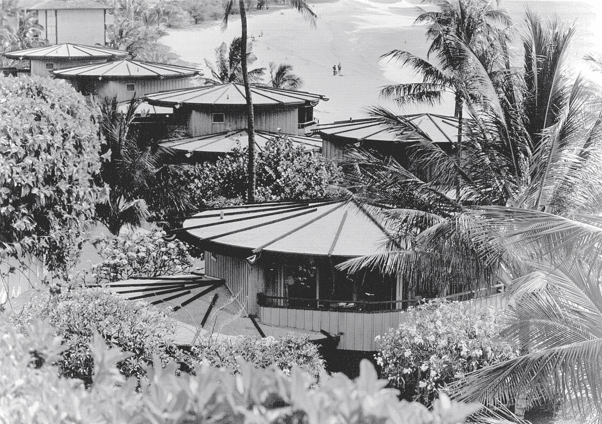 A historical black-and-white elevated photograph showing a cluster of circular, cottage-style resort cottages with fan-shaped roofs, surrounded by dense tropical foliage and palm trees near a sandy shore.