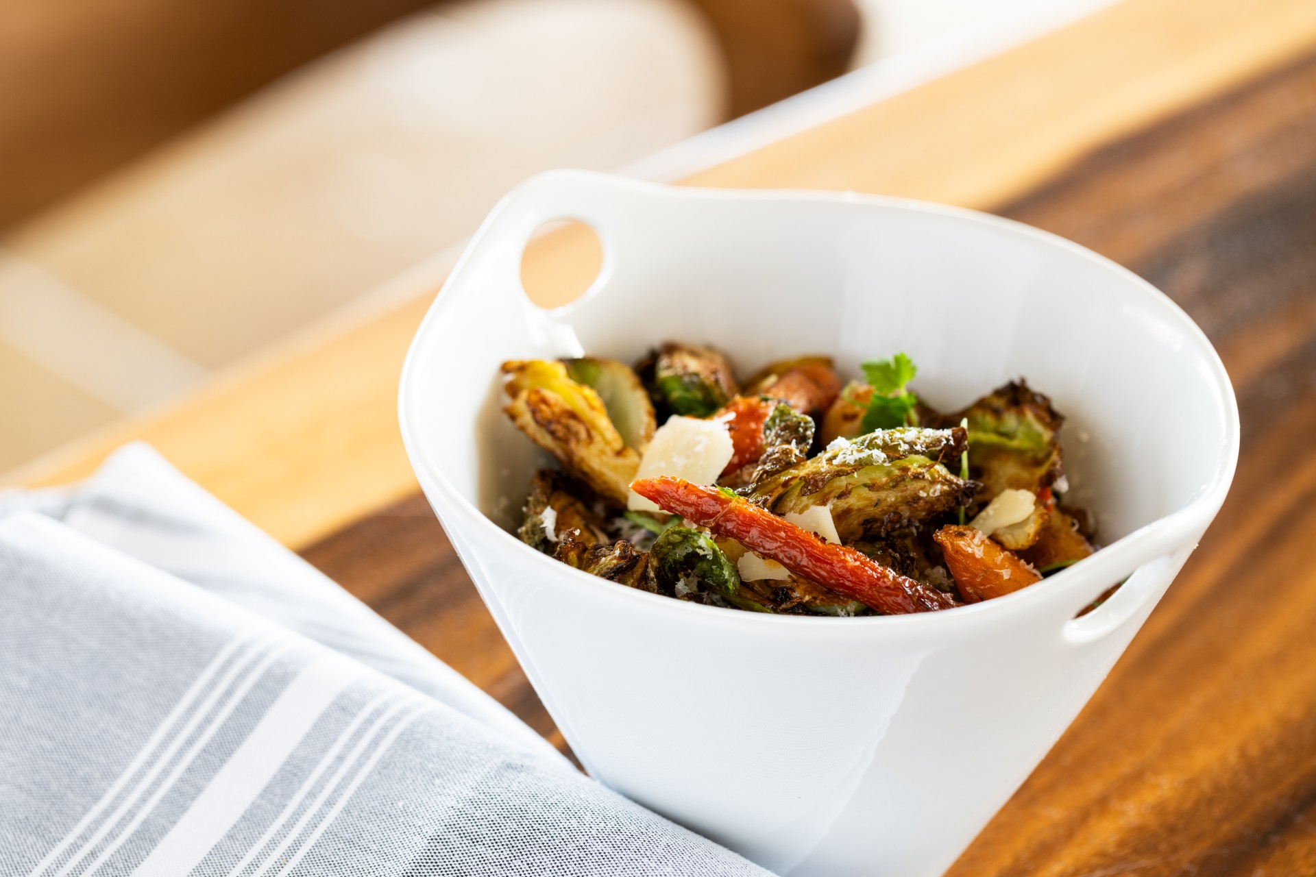 A white ceramic serving bowl filled with crispy roasted Brussels sprouts and glazed carrots, topped with shaved Parmesan cheese, resting on a wooden table beside a striped cloth napkin.