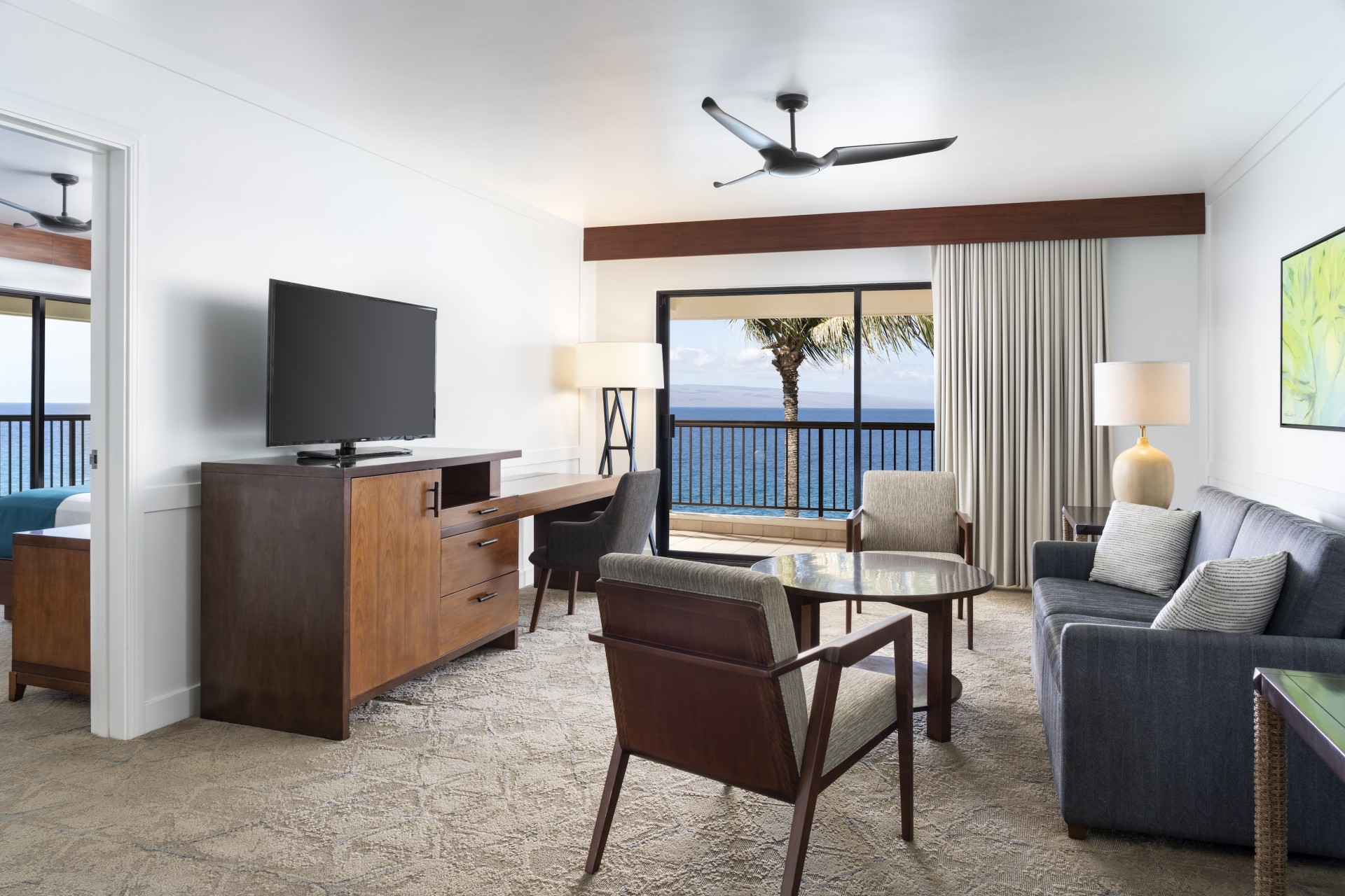 A oceanfront hotel suite living room featuring a dark grey sofa, wooden armchairs around a coffee table, a console table with a TV, and a balcony doorway opening to scenic ocean views.