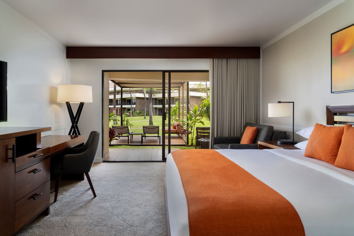 A resort guest room with vibrant orange bedding accents and pillows, dark wood furniture including a writing desk, and sliding glass doors opening onto a private patio with outdoor lounge seating and views of lush green lawn and tropical foli