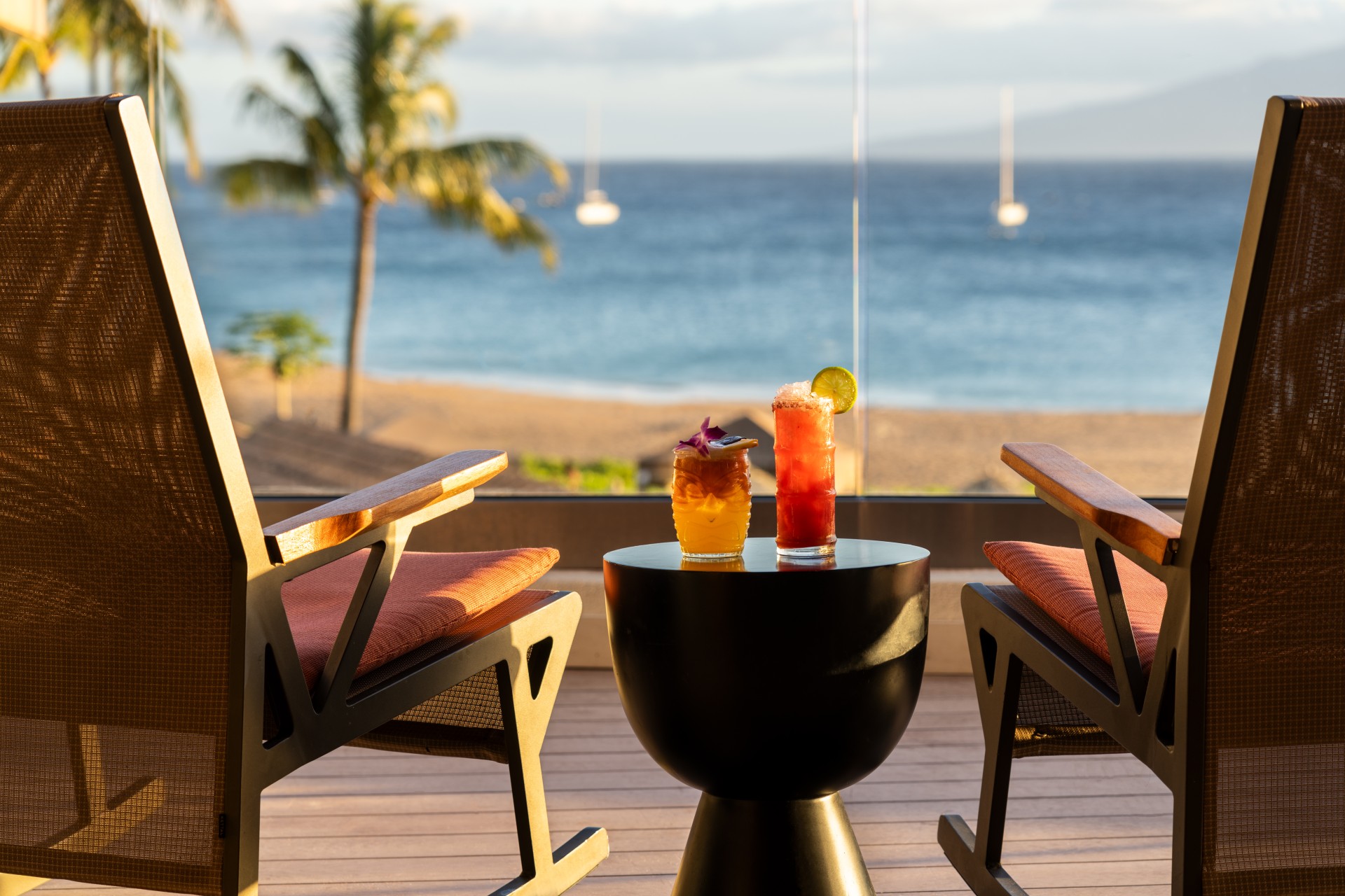 a table with drinks on it and chairs on a deck with a beach in the background