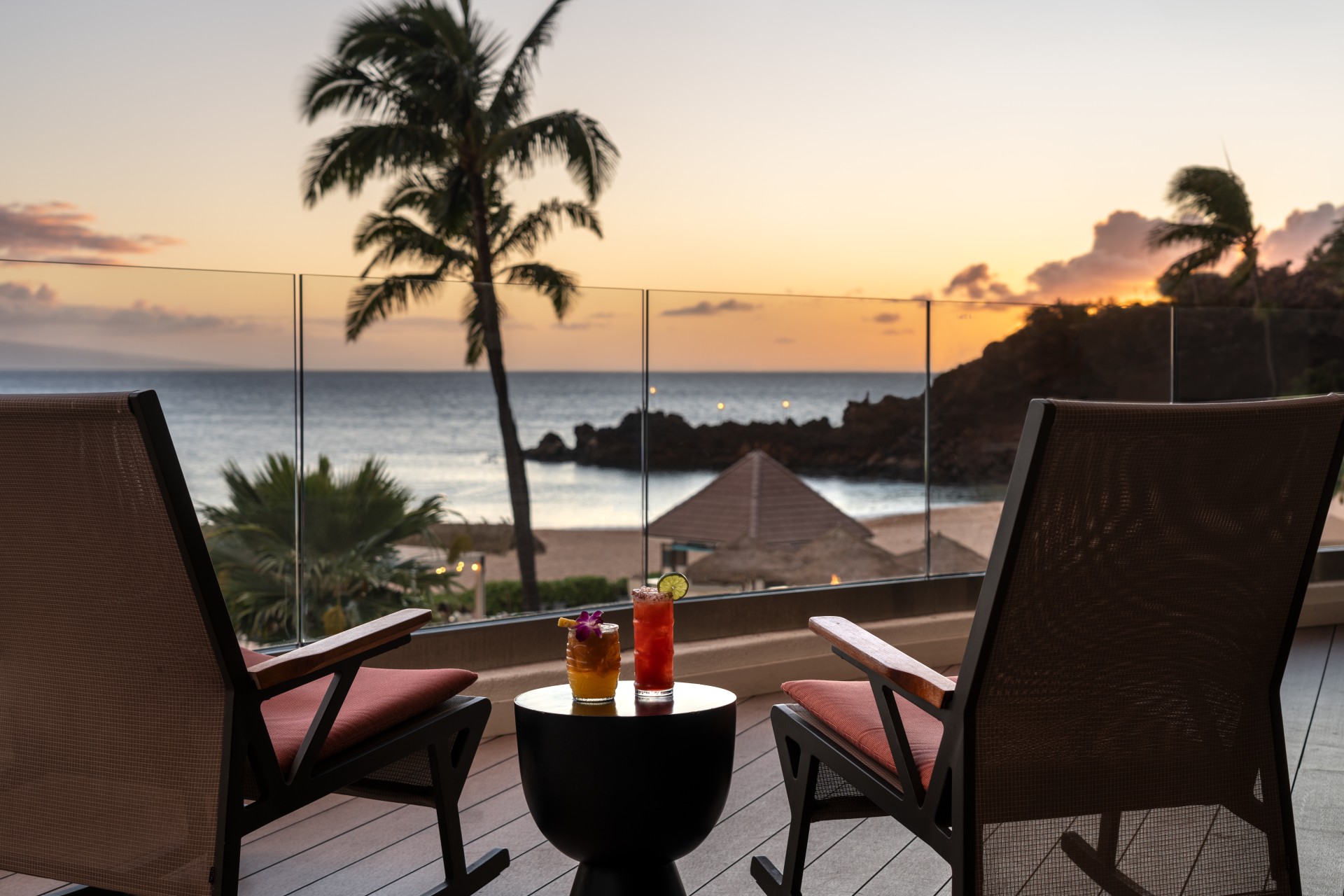 a table with drinks on it and chairs on a deck with a beach and water in the background