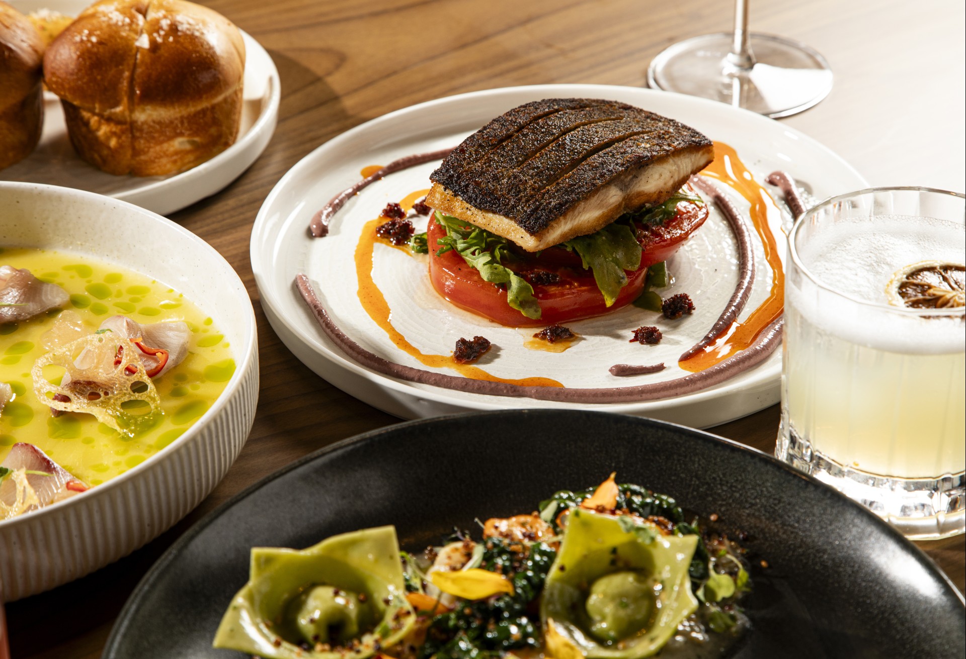 A gourmet dining table arrangement featuring crispy pan-seared fish stacked over sliced tomatoes, a bowl of yellow broth with fresh crudo, green ravioli pasta, fresh bread rolls, and a craft cocktail.