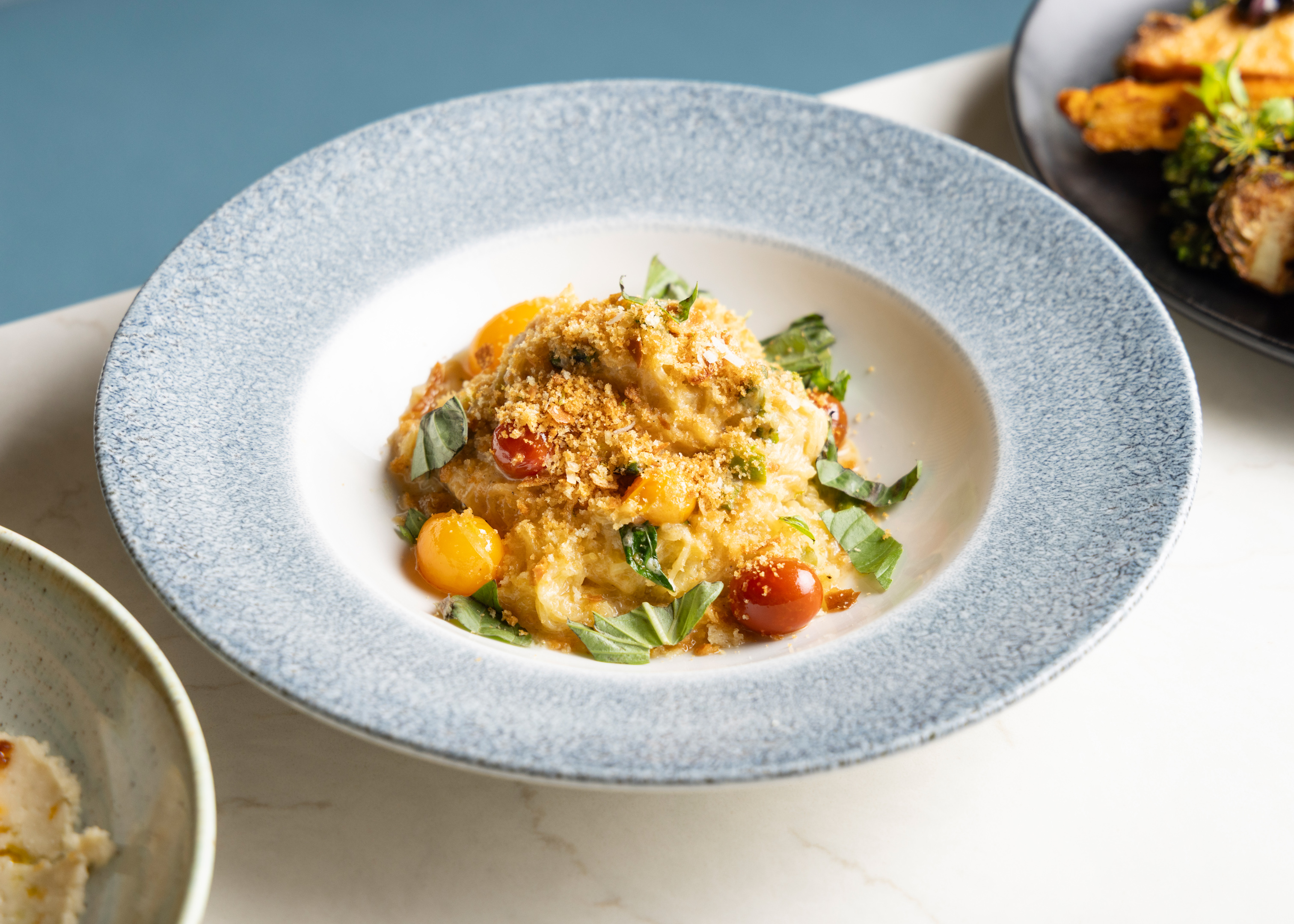 A shallow speckled blue bowl filled with a pasta dish topped with toasted breadcrumbs, whole red and yellow cherry tomatoes, and fresh torn basil leaves.