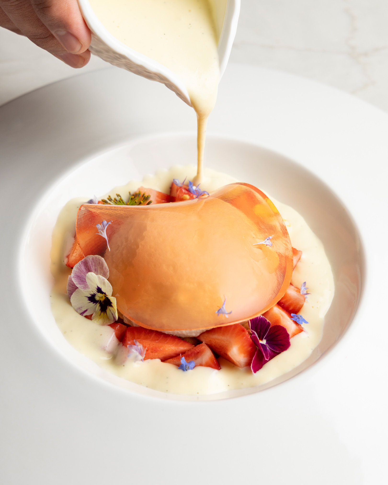 A hand pouring a rich white sauce over an elegant gourmet dessert featuring fresh sliced strawberries, delicate edible flowers, and a translucent orange dome in a white shallow bowl.