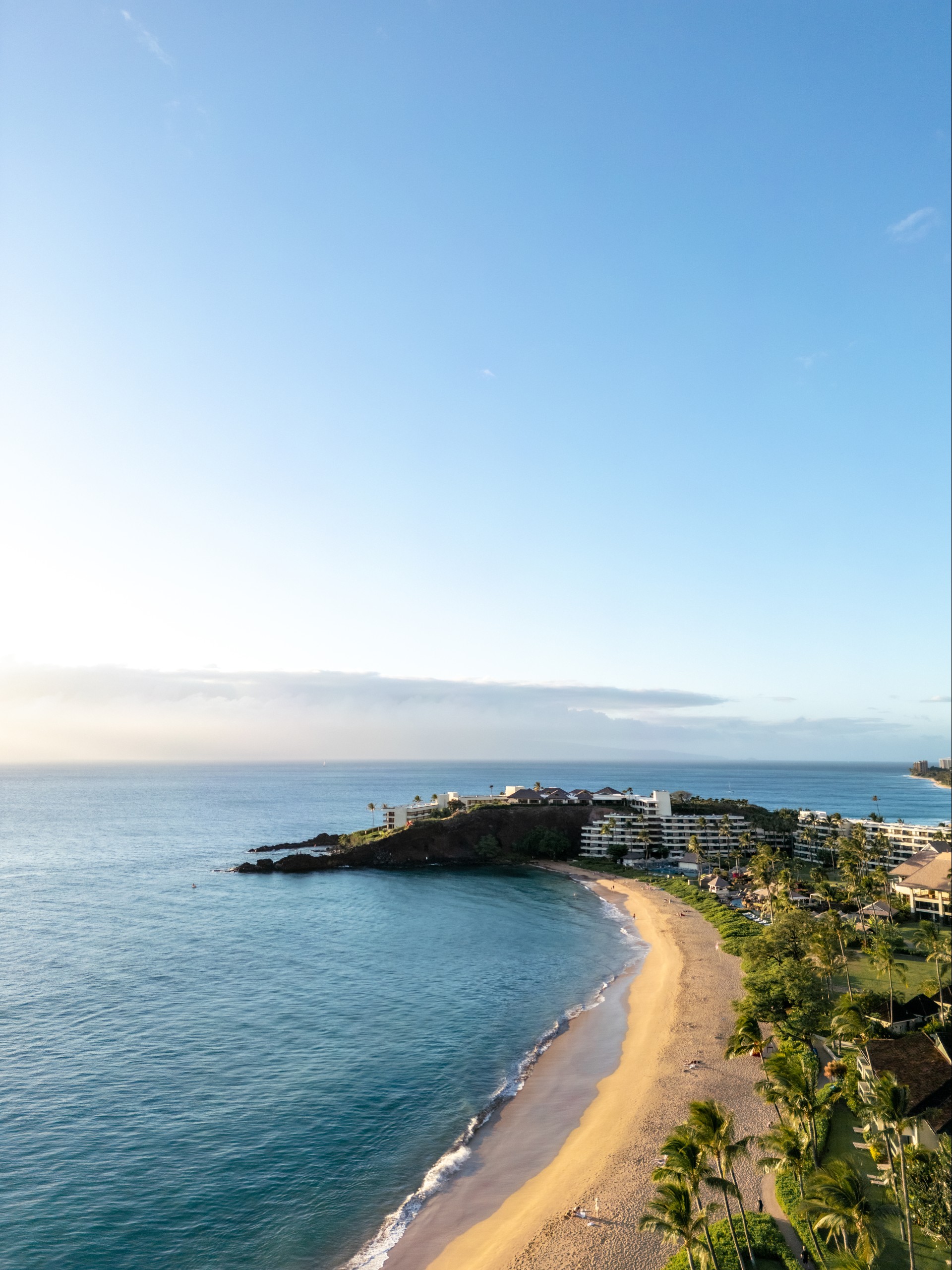 A high-angle vertical landscape view of a golden sandy beach curving along deep blue ocean waters toward a rocky promontory with resort buildings, under a clear sky bathed in soft sunlight.