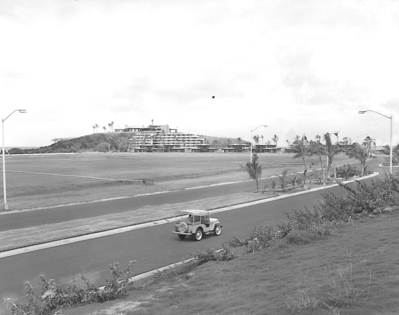 historical black-and-white photograph showing a small vintage utility vehicle driving down a newly paved road, with open land, palm trees, and coastal resort structures in the background.