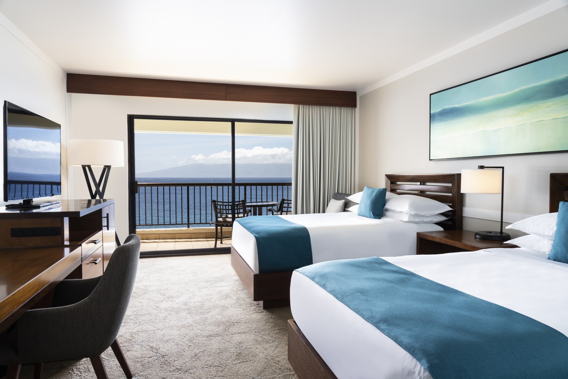 A modern resort guest room with two beds accented by teal runners and pillows, dark wood furniture, coastal artwork, and sliding glass doors opening to a balcony with panoramic ocean views.