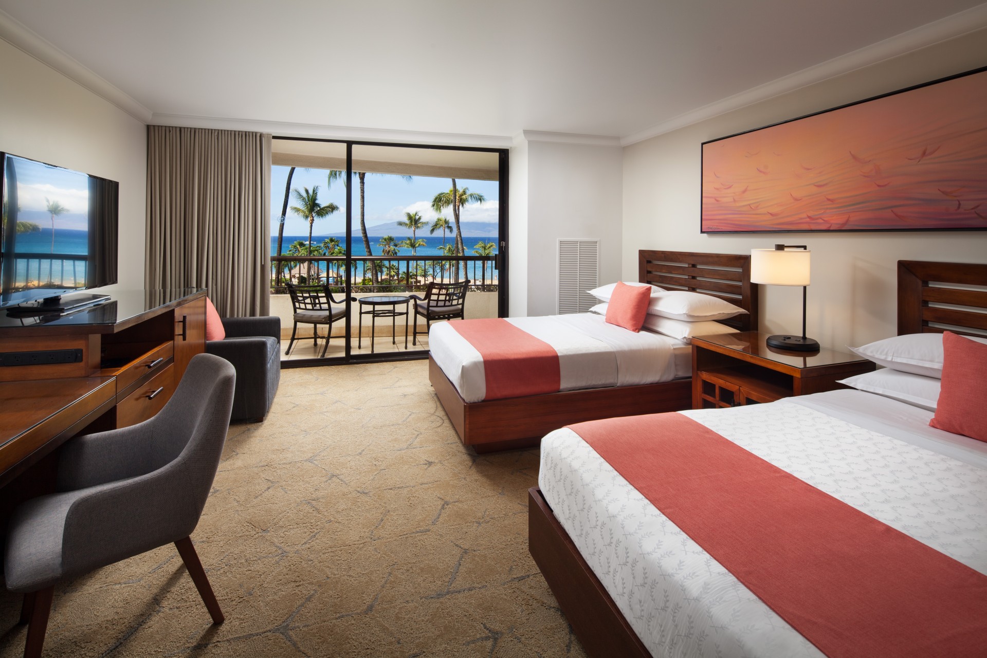 A hotel guest room with two double beds featuring coral-red runners and pillows, dark wood furnishings, a desk chair, and sliding glass doors that lead to a private balcony with seating and ocean views.