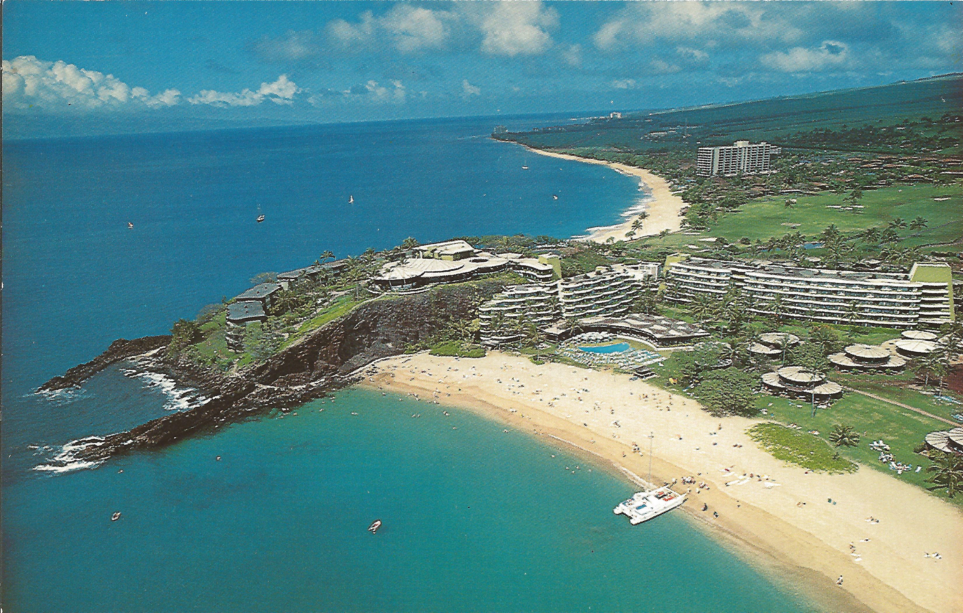 A vintage postcard aerial view showing a bustling beach resort along a clear turquoise bay, featuring a sandy beach with a catamaran, rocky cliffs, swimming pools, and surrounding green landscapes.