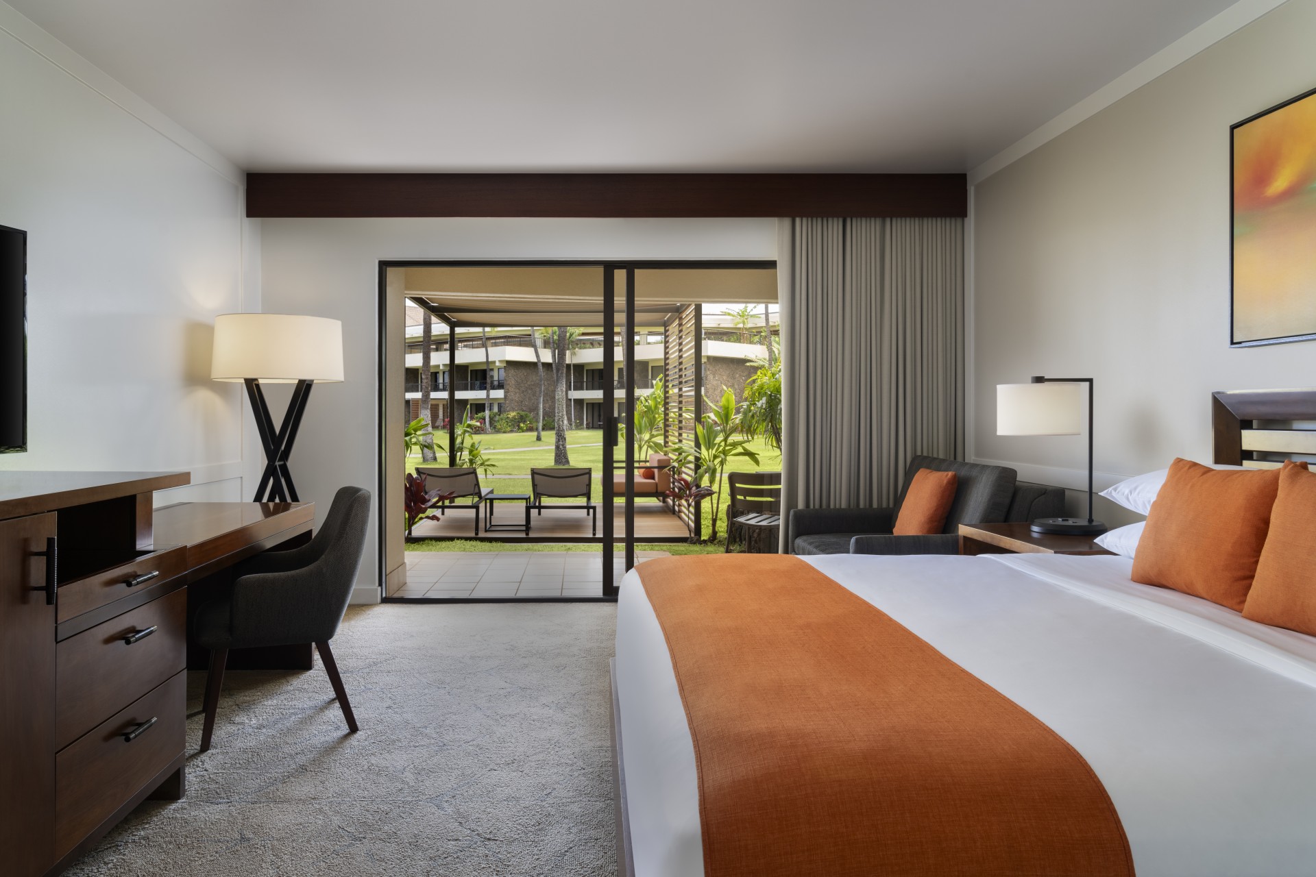 A ground-floor resort guest room with vibrant orange bedding accents and pillows, dark wood furniture including a writing desk, and sliding glass doors opening onto a private patio with outdoor lounge seating and views of lush green lawn and tropical foli