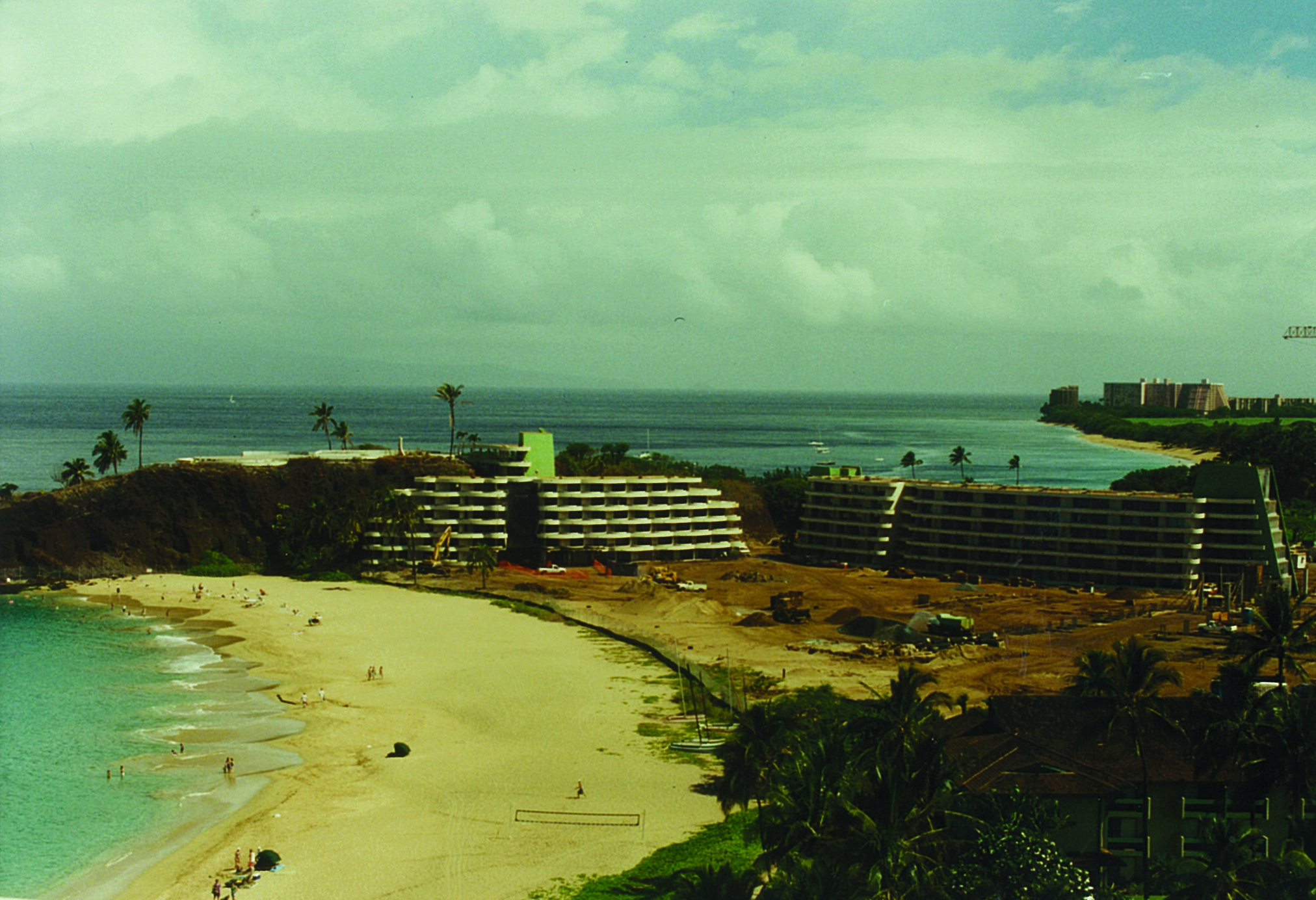 A vintage photograph capturing the construction phase of a coastal hotel resort situated along a sandy beach, featuring multi-story building frames and earth-moving equipment set against an expanse of ocean.