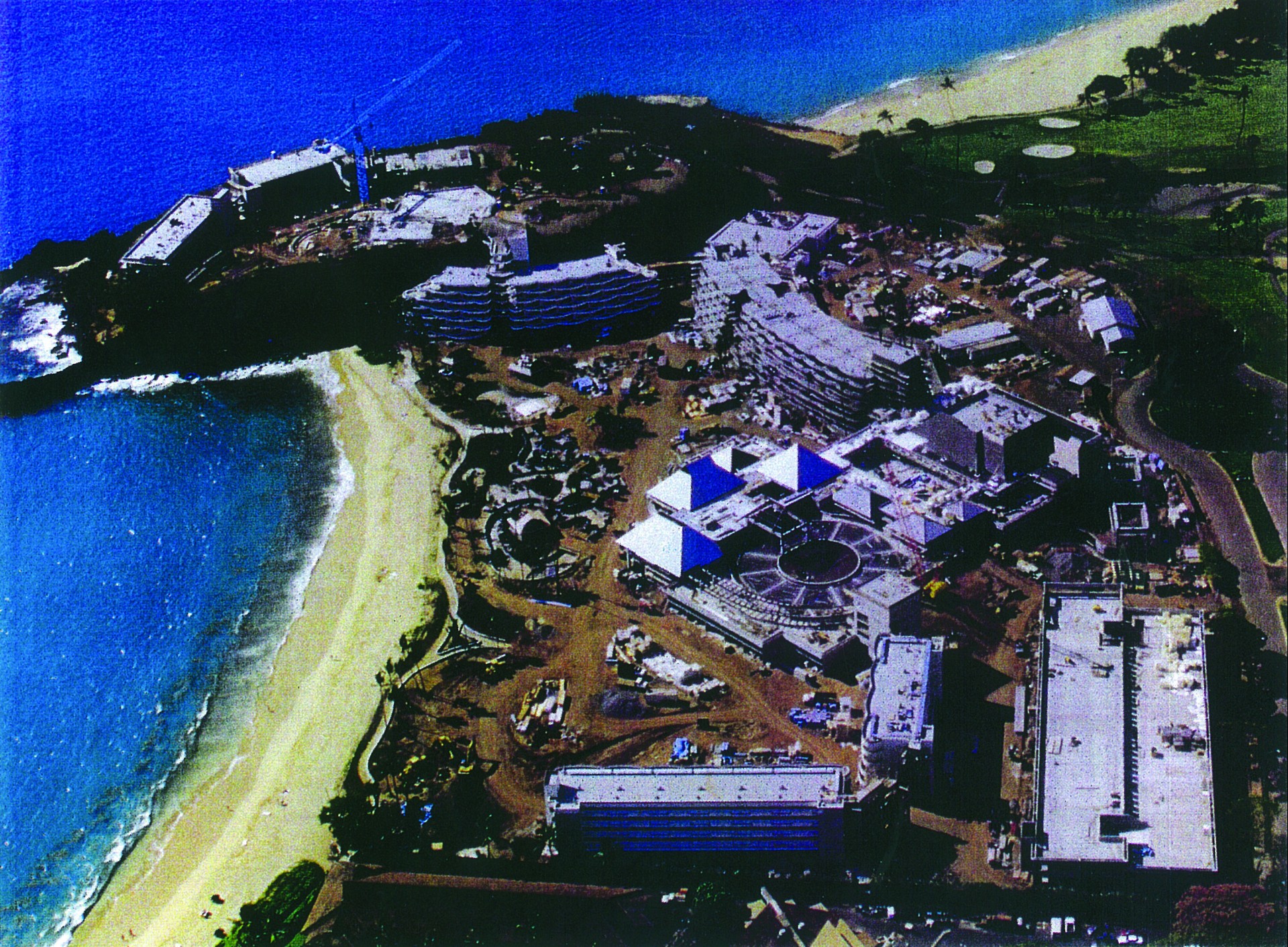 An aerial view showing the construction site of a large beachfront resort complex situated along a sandy cove next to turquoise ocean water.