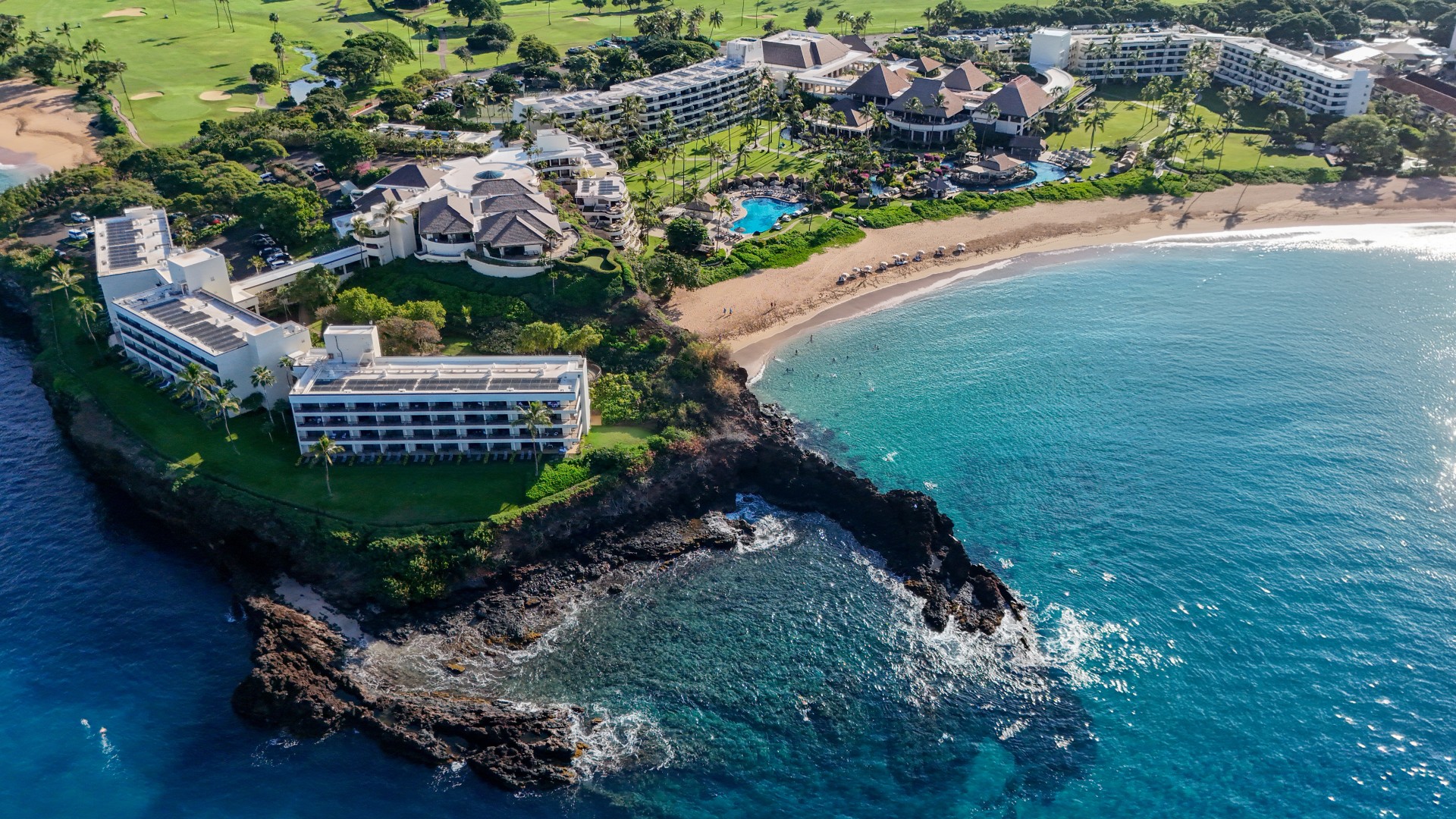 A high-angle aerial view of a seaside resort complex built along a dramatic dark lava rock point, featuring oceanfront hotel wings, swimming pools, a sandy beach, and surrounding green golf courses bordering bright blue water.