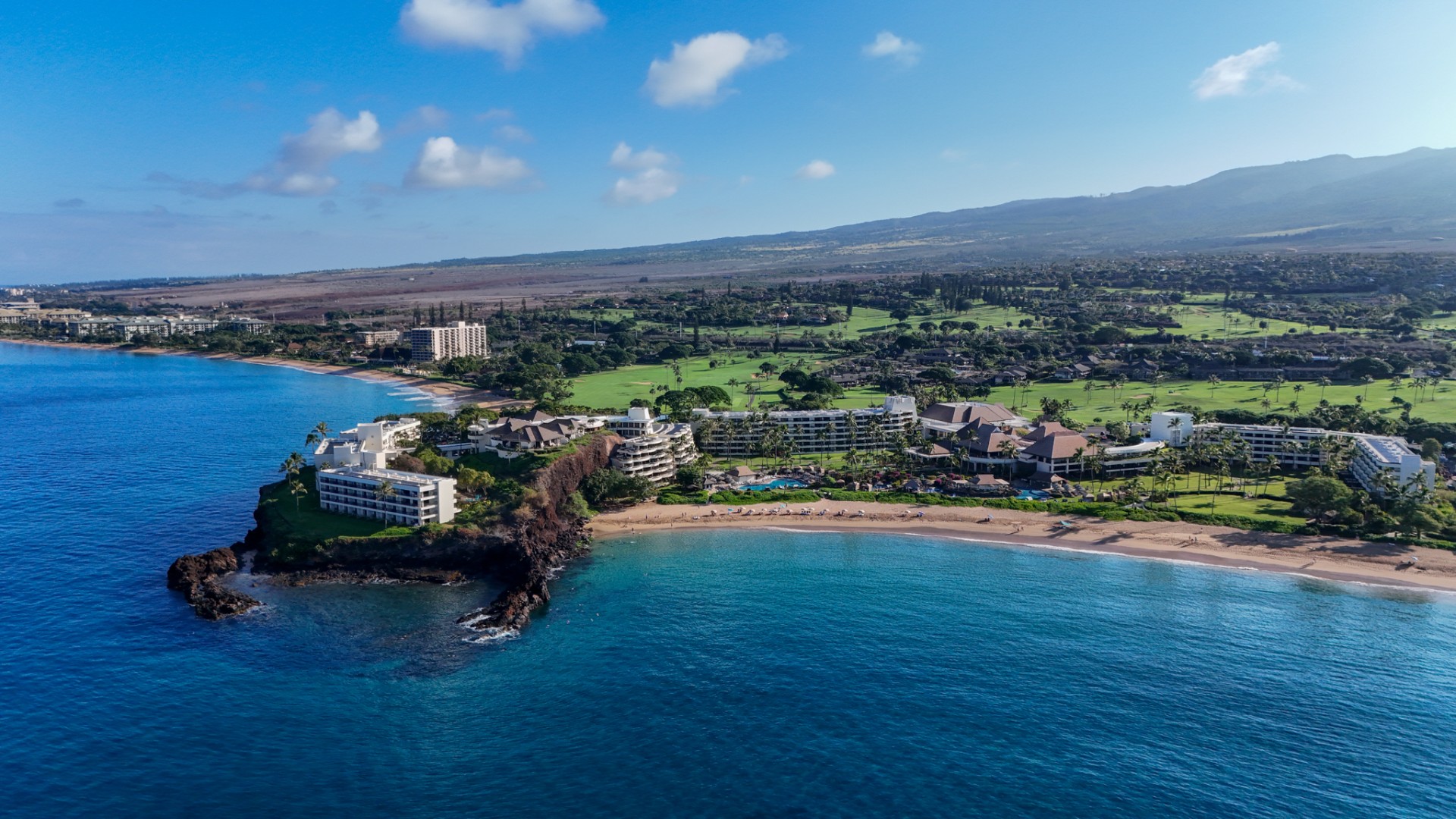 A wide aerial view of a coastal tropical resort nestled along a sandy cove and rocky point, with vibrant turquoise ocean waters in the foreground and a golf course against mountains in the background.
