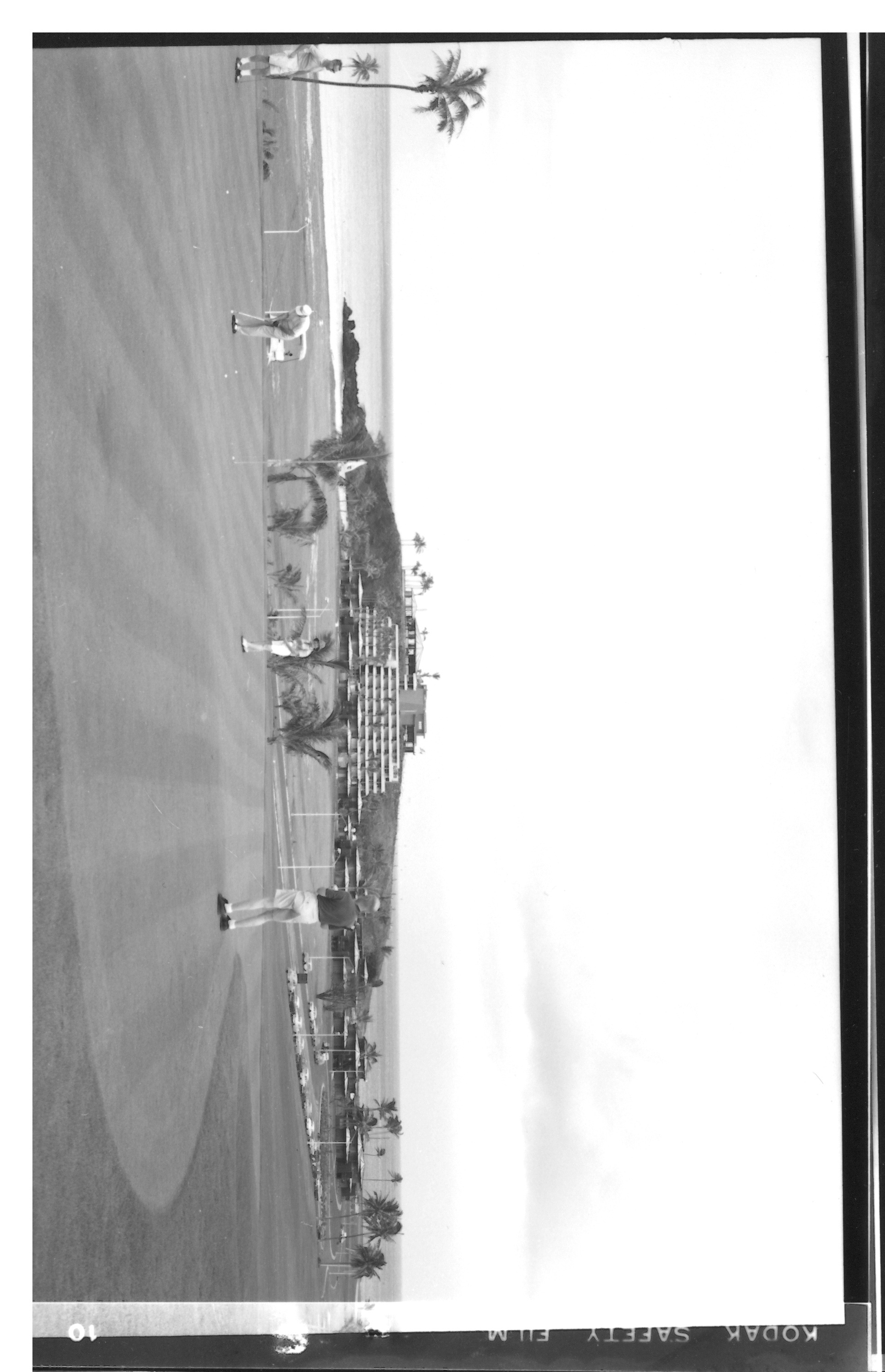 A historical black-and-white film frame showing golfers practicing on a green with a flagstick, overlooking a coastal resort complex built into a hillside near the ocean.