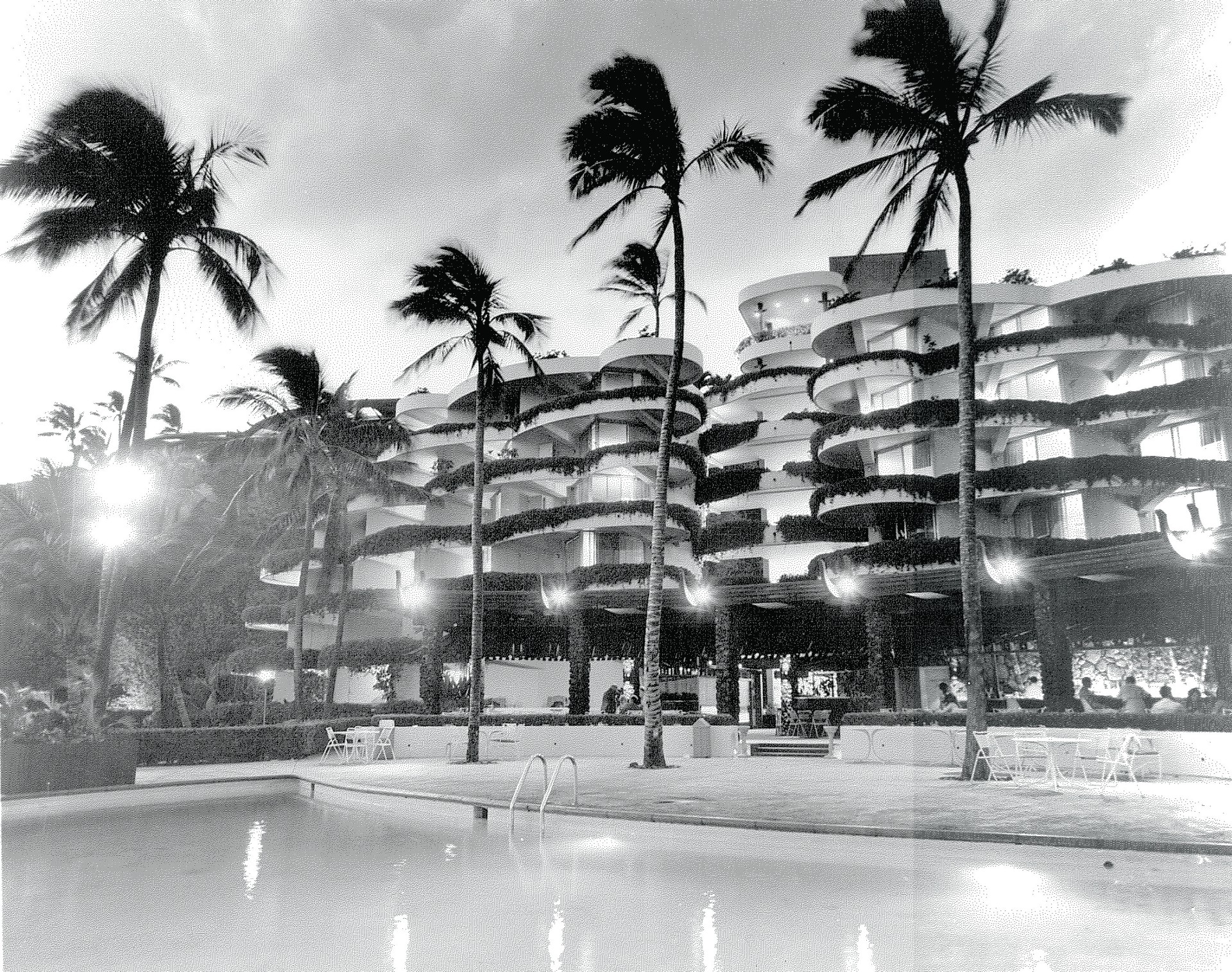 A historical black-and-white night photograph showing a illuminated resort pool patio framed by tall palm trees, in front of a multi-story hotel building with curved, vine-draped balconies.