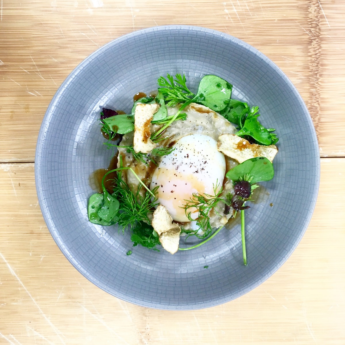 A top-down view of a gourmet dish served in a light grey textured bowl, featuring a soft-poached egg, fresh green herbs, croutons, and a drizzle of jus over a creamy puree, set against a wooden surface.