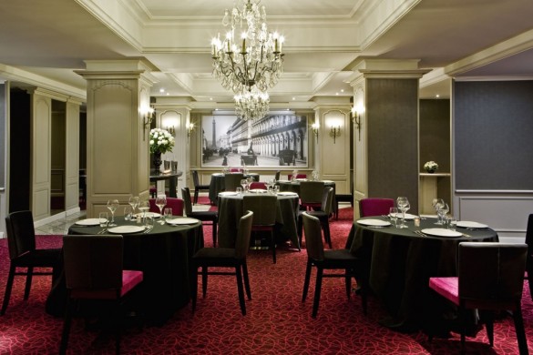 A sophisticated dining room featuring dark grey tableclothed round tables, a crystal chandelier, magenta patterned carpet, dark and magenta upholstered dining chairs, and a large black-and-white historic cityscape photograph framed on the back wall.