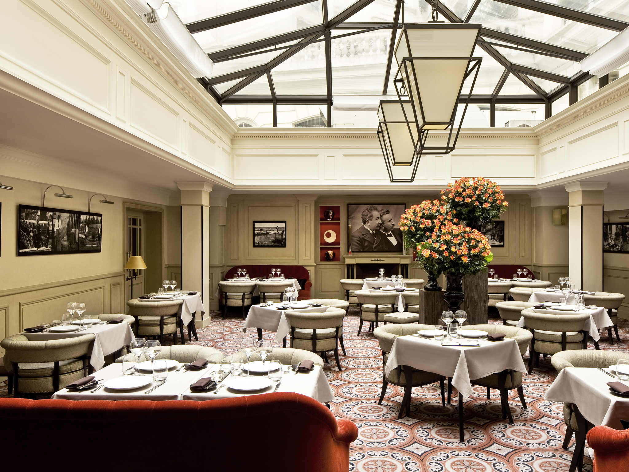An elegant fine dining restaurant featuring a large glass skylight ceiling, suspended lantern chandeliers, white-clothed tables, beige tub armchairs, red velvet banquettes, patterned tiled flooring, and a large central orange flower arrangement.