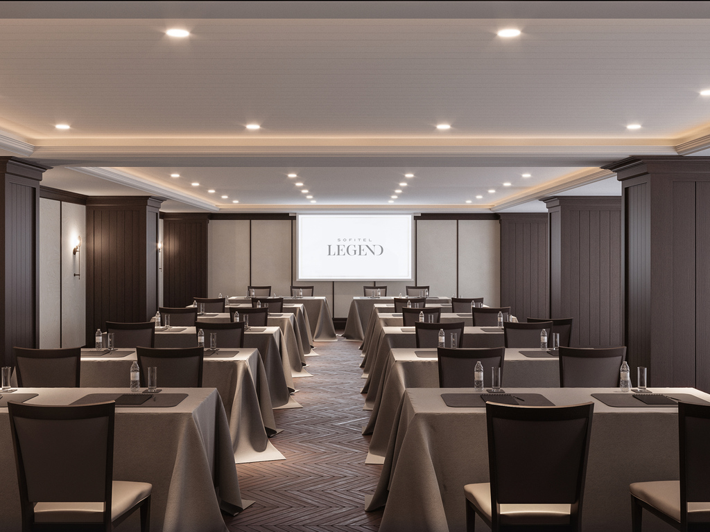 Modern conference room with classroom-style setup, dark wood-paneled walls, grey-draped tables with chairs, a central aisle, and a projection screen displaying 'SOFITEL LEGEND' at the front.