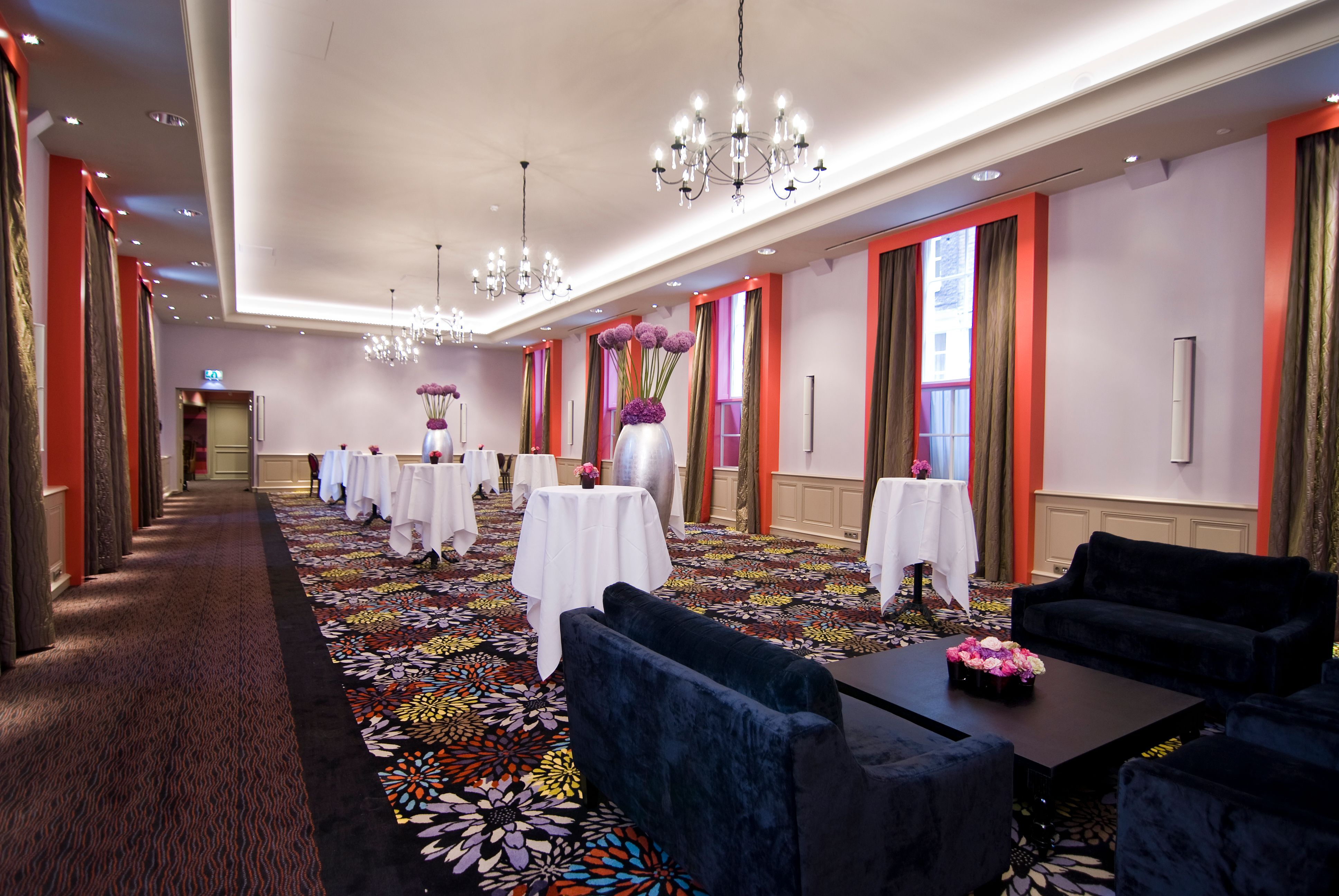 A grand event hall setup with high cocktail tables draped in white cloths, dark velvet lounge seating in the foreground, vibrant floral pattern carpeting, and bright orange accents framing large windows.