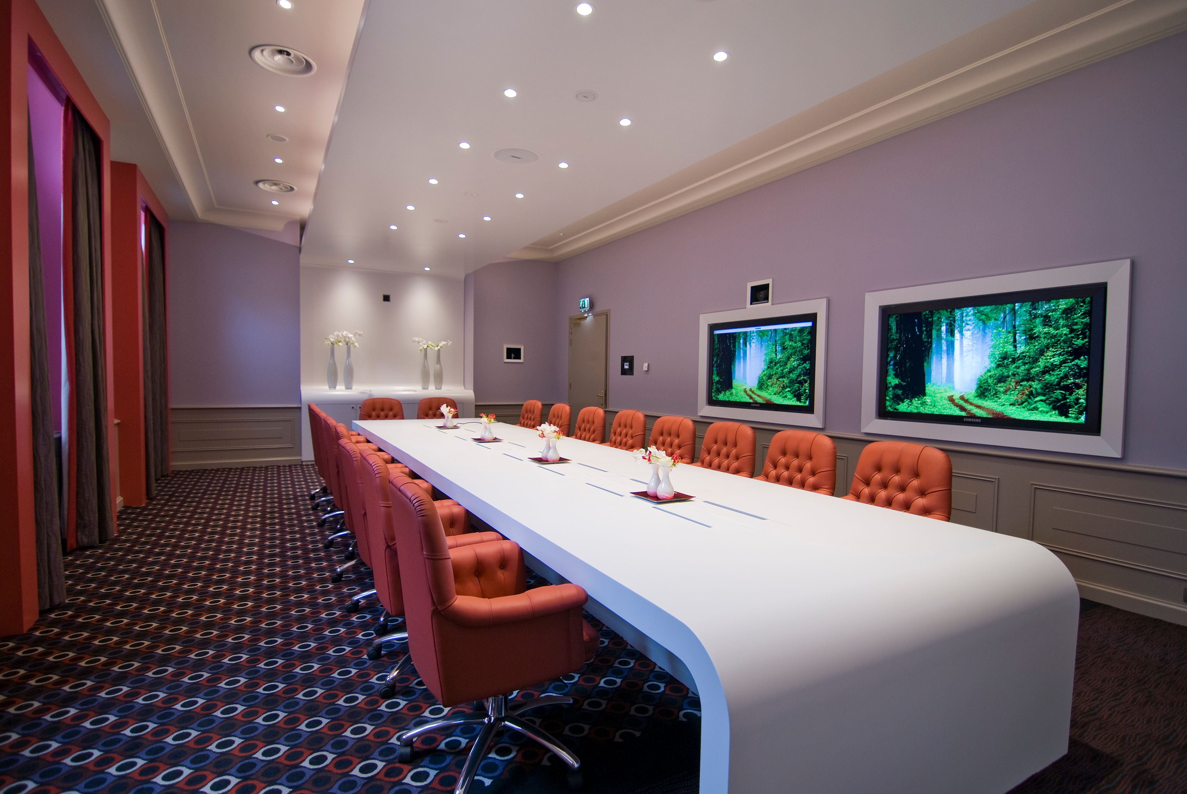 a long white table with orange chairs in a room with a television on the wall