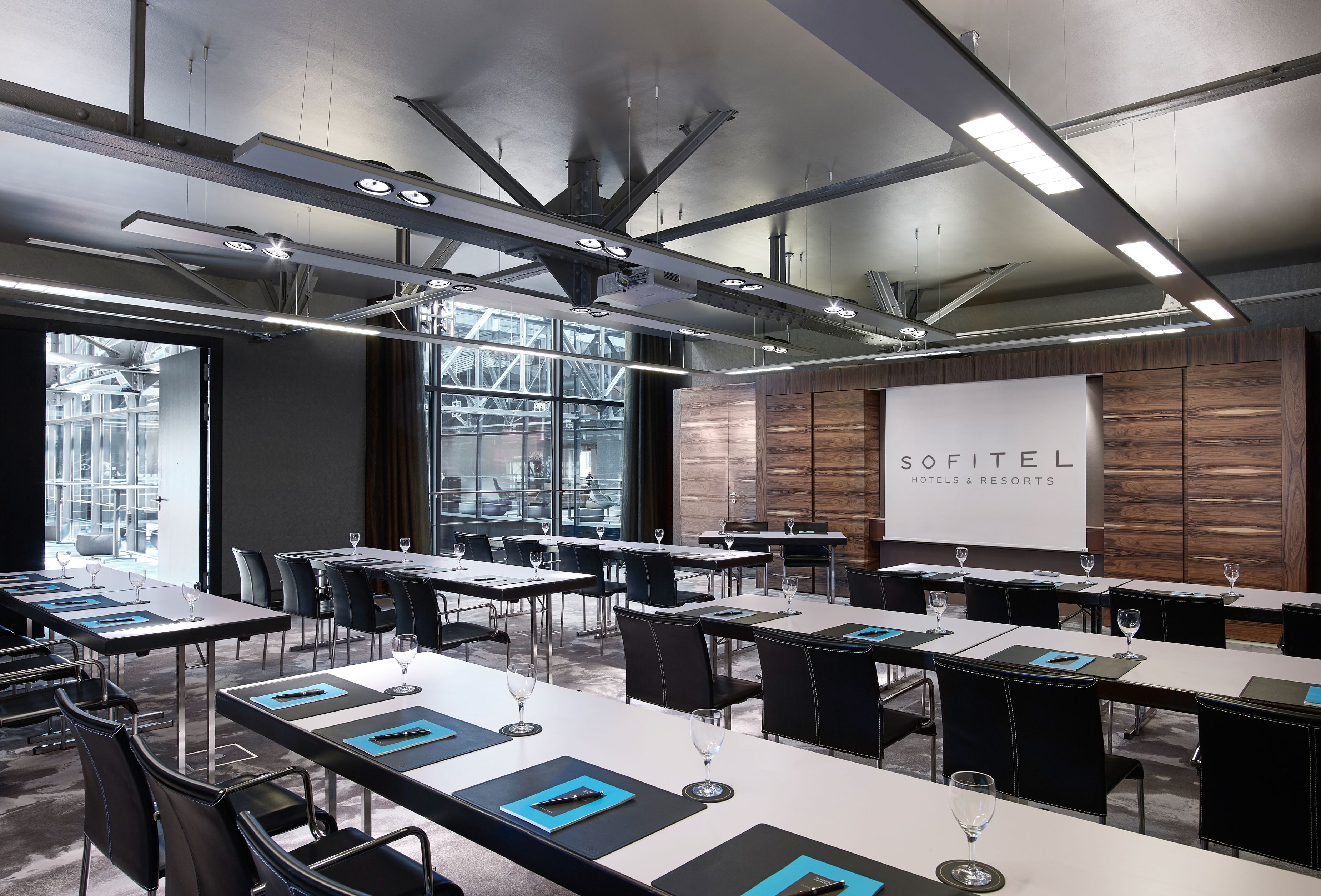 A modern, industrial-style conference room at Sofitel Hotels & Resorts, featuring long white training tables with black leather executive chairs, dark wood wall paneling around a large projection screen, exposed steel ceiling trusses with sleek track ligh