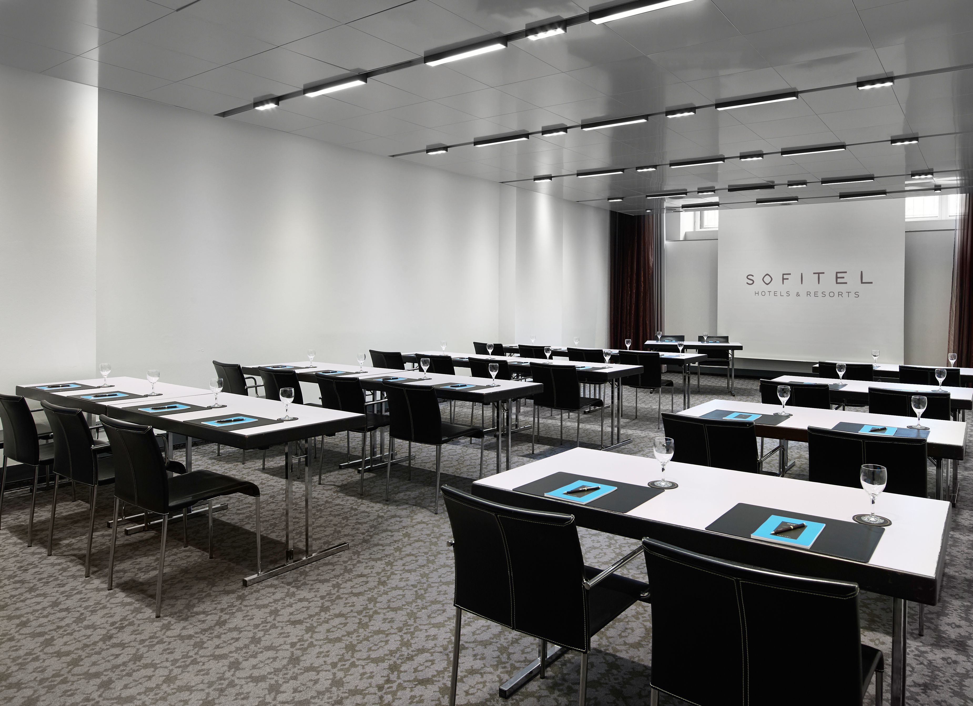 A sleek, professional meeting room set up classroom-style at Sofitel Hotels & Resorts, featuring long white tables set with note pads and water glasses, black leather chairs, modern linear track lighting across the ceiling, light patterned carpeting, and 