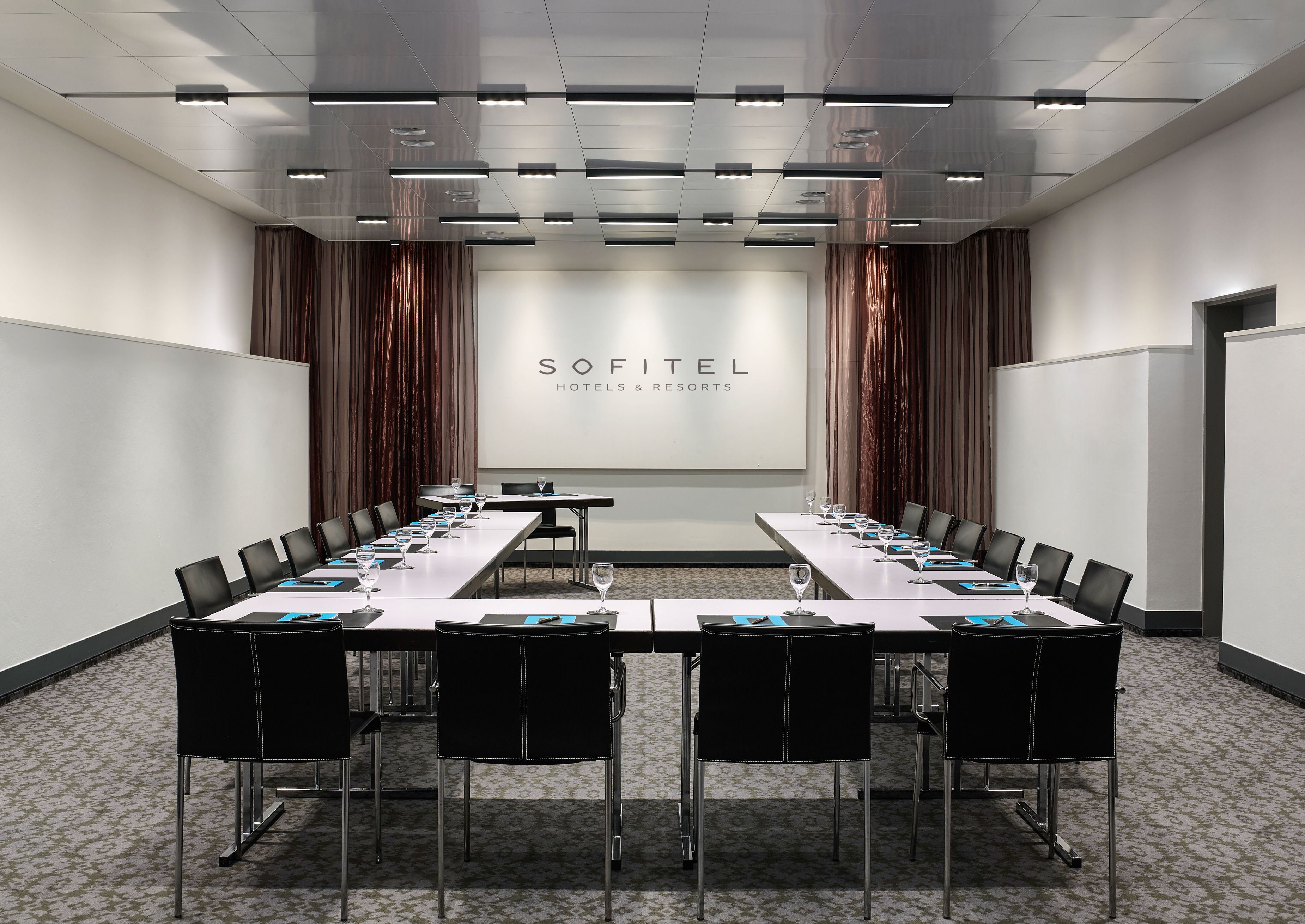 A modern, u-shaped meeting room configuration at Sofitel Hotels & Resorts, featuring long white tables set with black placemats and glassware, black leather armchairs, grey patterned carpet, linear track lighting on a reflective metallic ceiling, and sati
