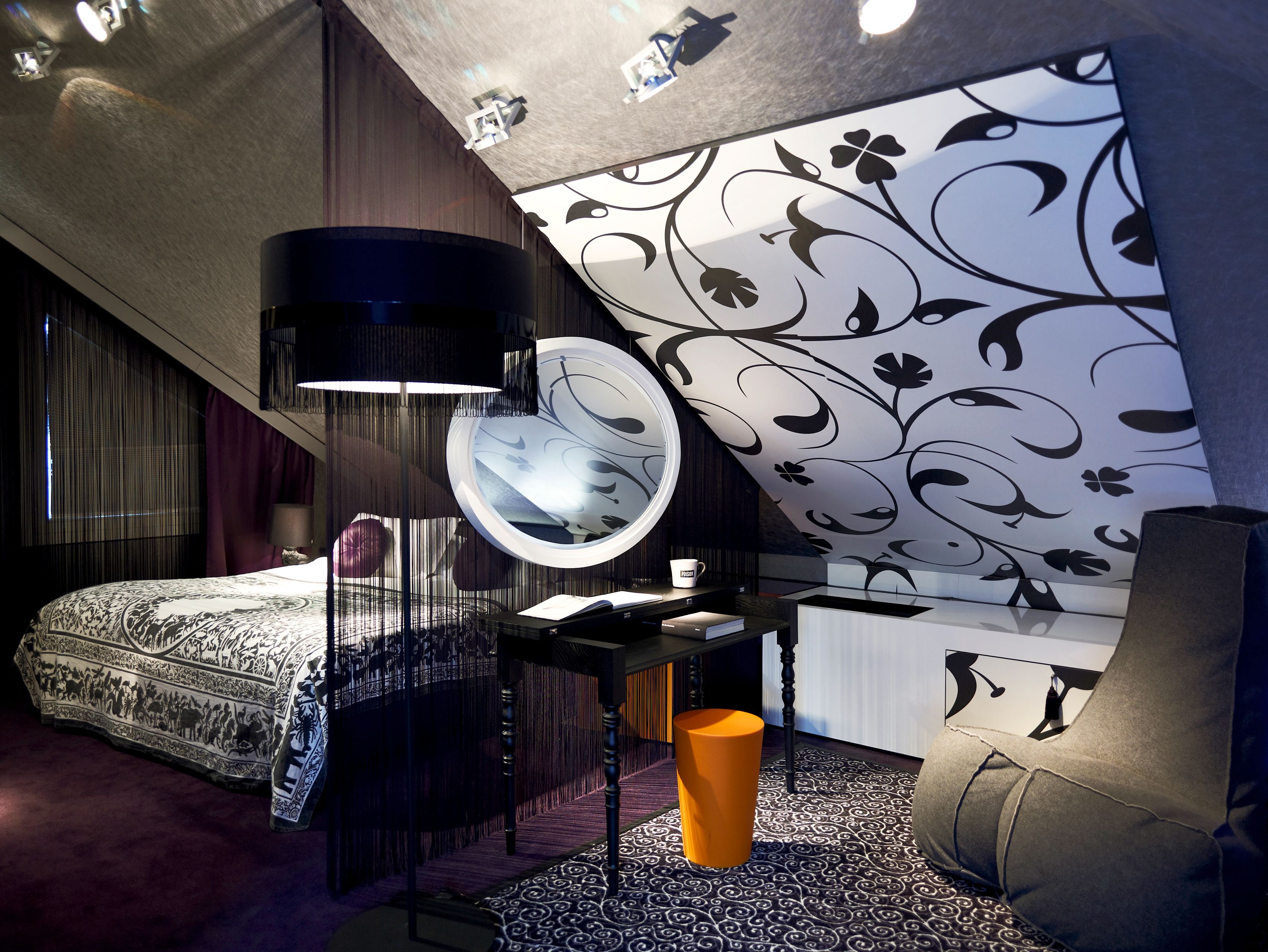 A stylish modern attic bedroom suite featuring sloped ceilings, bold black-and-white floral patterned wall panels, a black vanity table with a round mirror, an orange cylindrical stool, a black floor lamp with fringe, and a bed dressed in an intricate pat