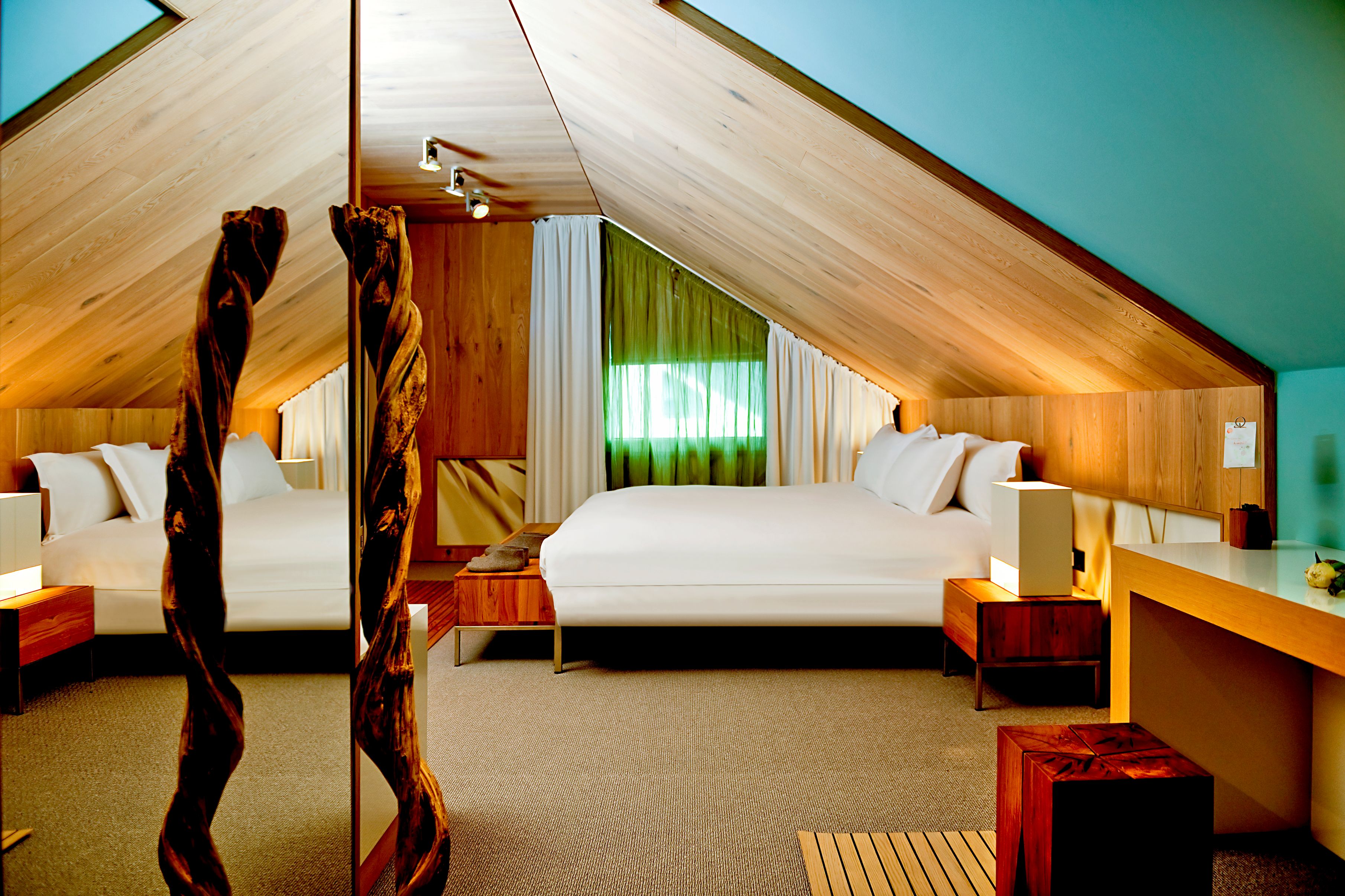 A modern attic bedroom featuring light wood-paneled sloped ceilings, a crisp white bed, turquoise wall accent, a large floor-to-ceiling mirror framed by carved twisted wooden posts, and warm bedside lighting.