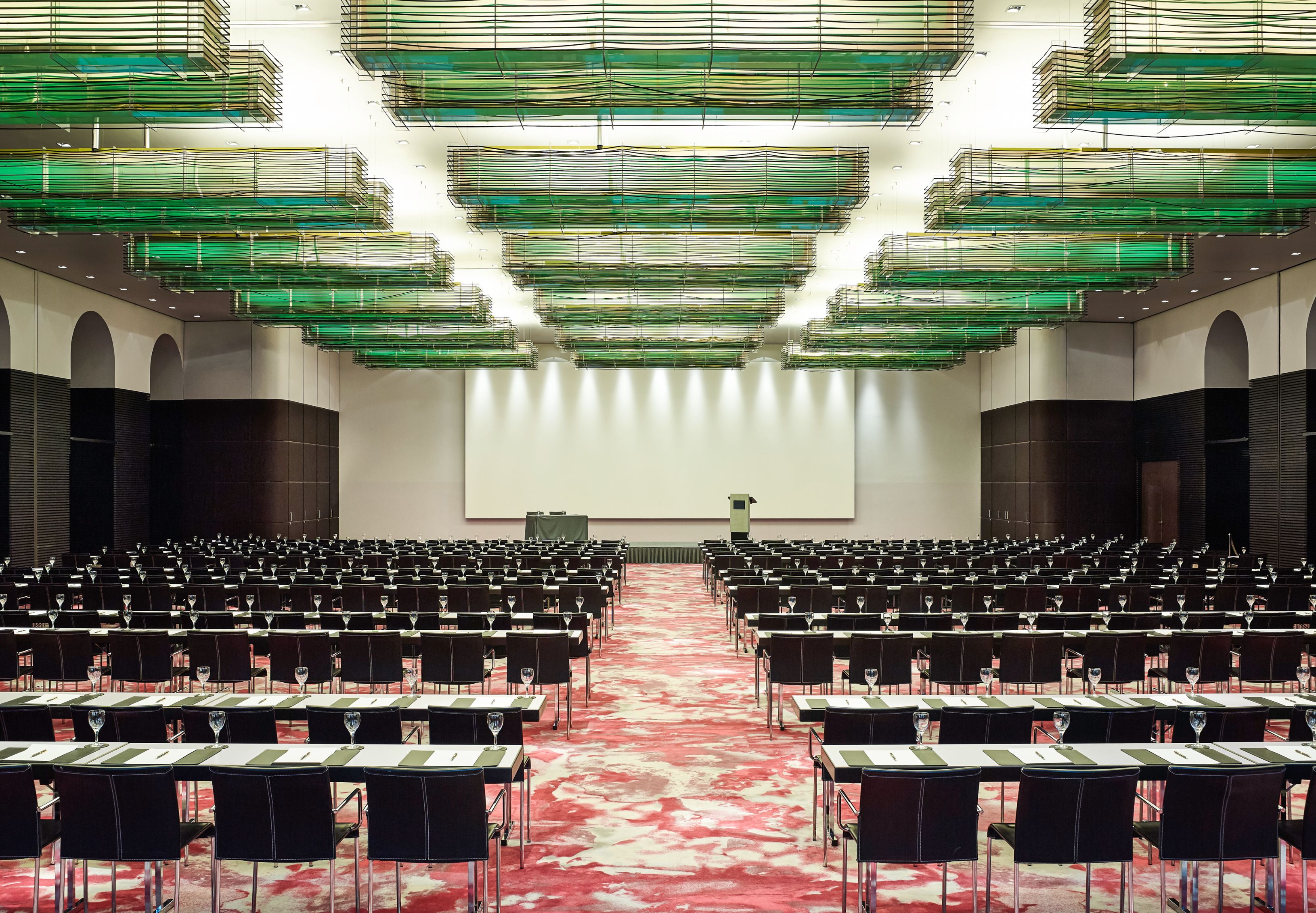 A grand, spacious conference hall configured in classroom seating, featuring long rows of dark tables and black chairs over a vibrant pink and white patterned carpet, a large white presentation screen at the front, and impressive green layered ceiling lig