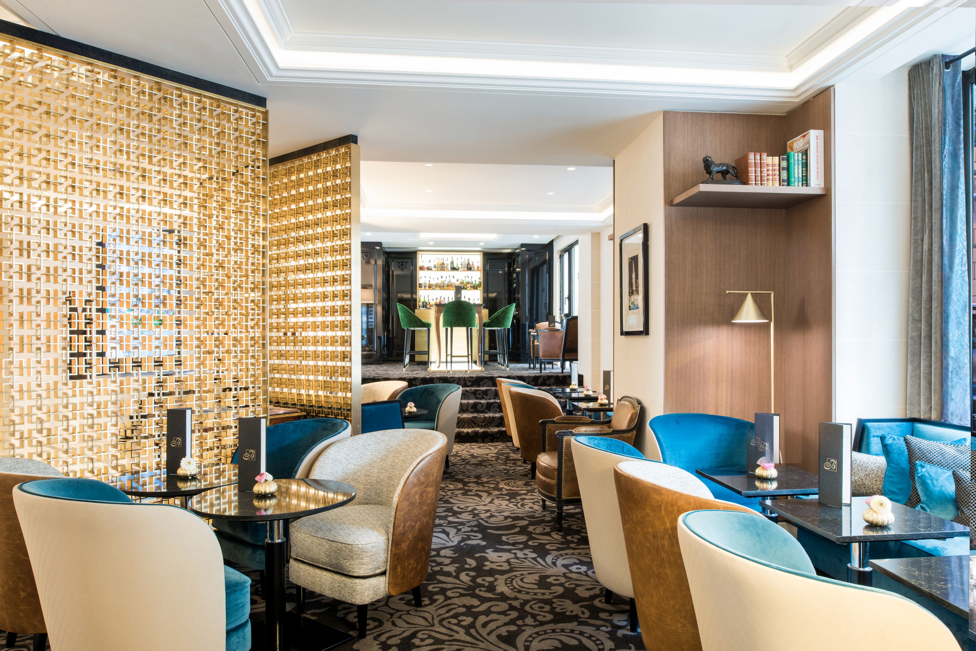 A stylish hotel bar lounge featuring plush velvet armchairs in teal, cream, and brown, decorative brass geometric room dividers, small bistro tables, and an illuminated bar counter visible in the background.