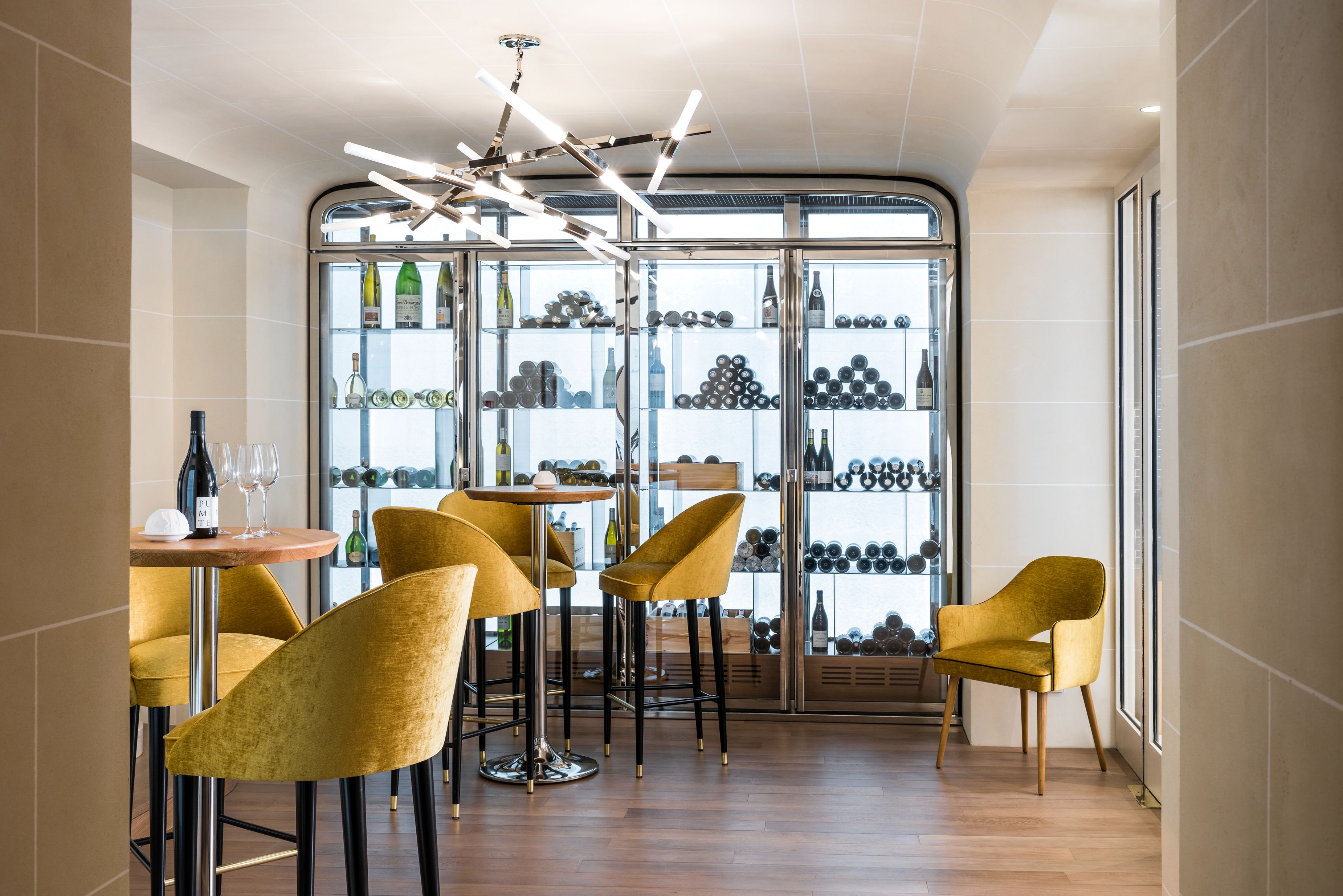 A modern wine tasting room featuring mustard-yellow velvet bar stools set around tall bistro tables, light wood flooring, a sputnik-style tubular light fixture, and a large illuminated floor-to-ceiling glass wine refrigerator.