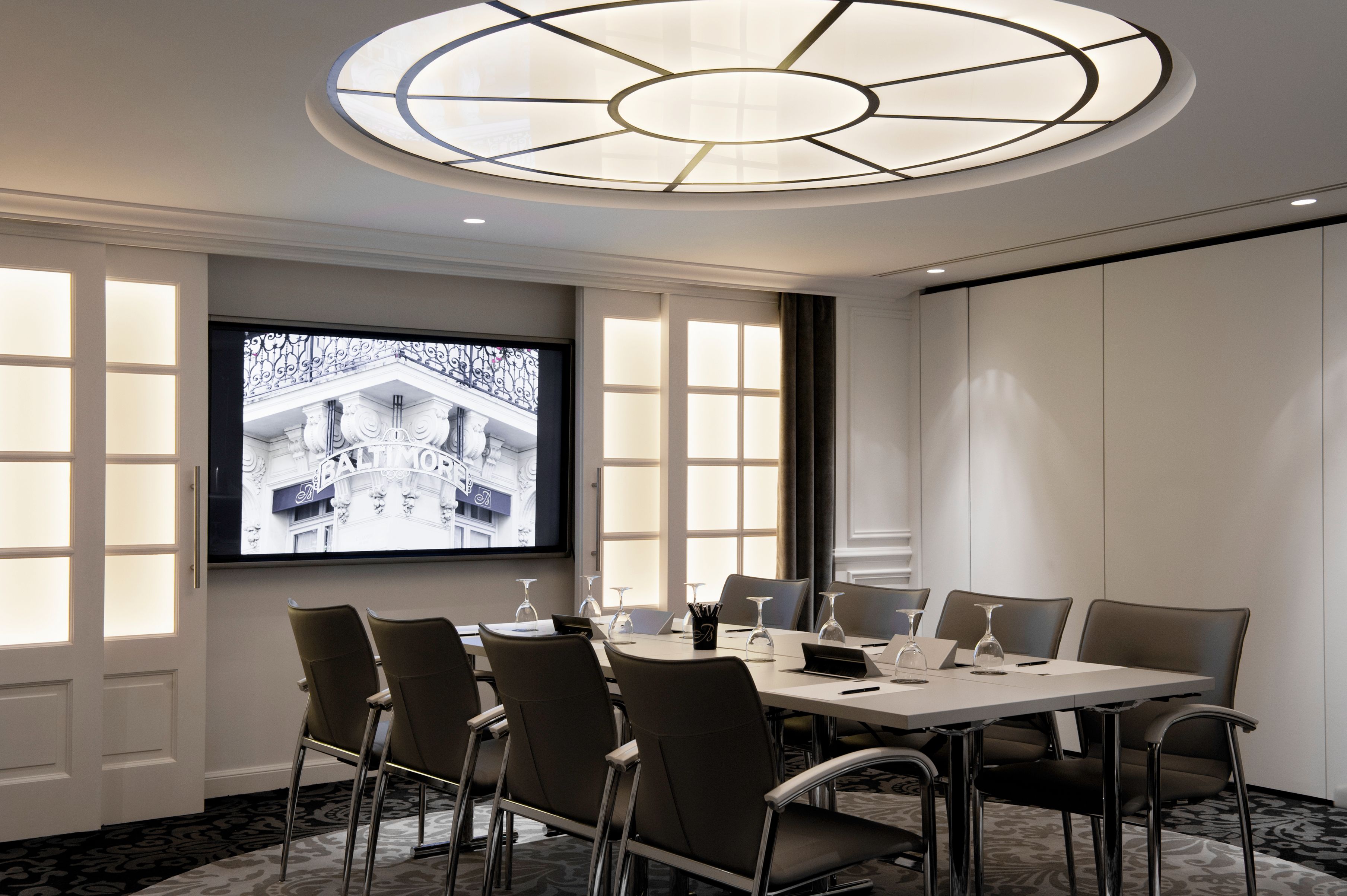 Modern conference room set with a long grey boardroom table surrounded by executive chairs, featuring a circular geometric ceiling light fixture and a wall-mounted flat-screen television displaying an architectural image.