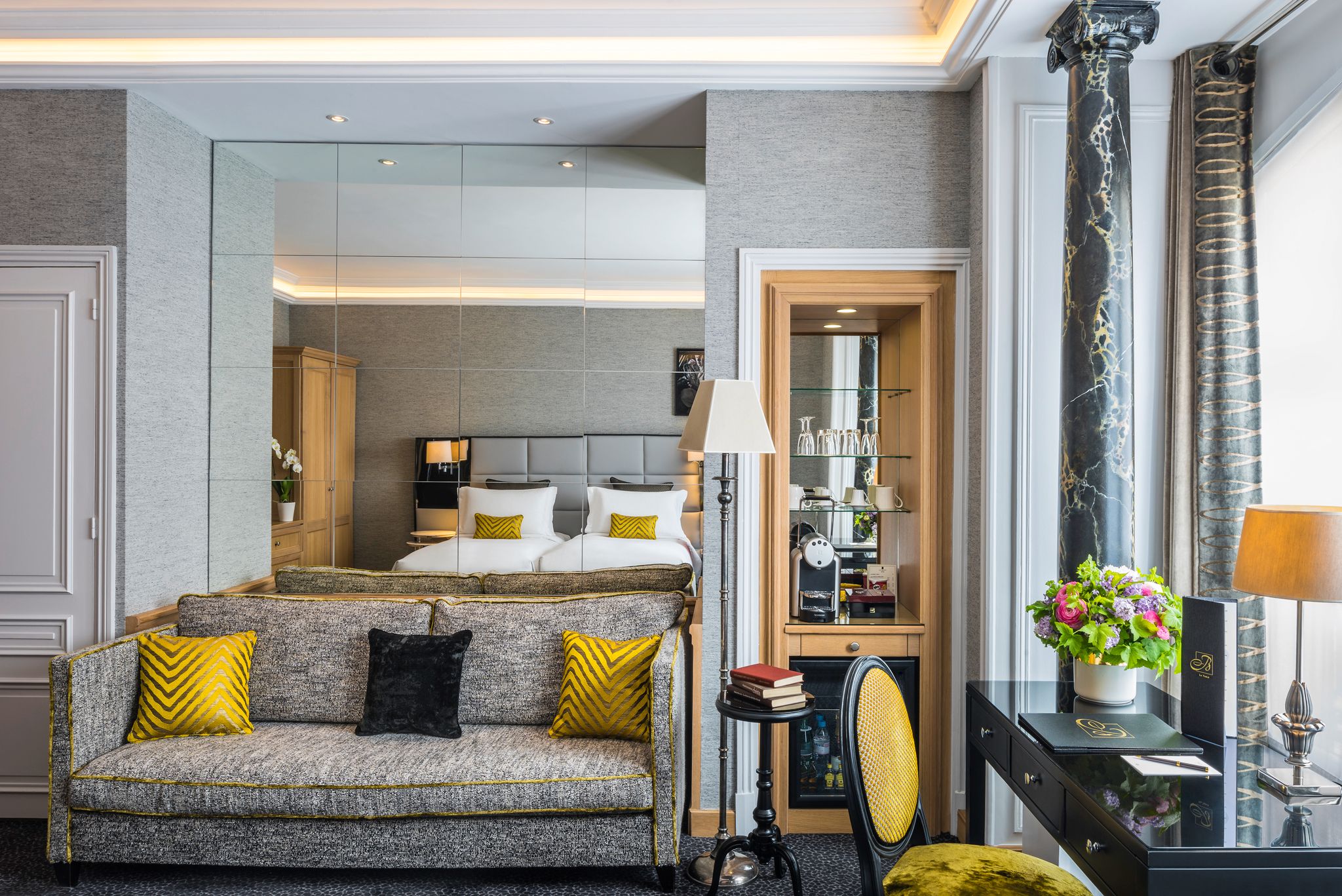 A stylish hotel suite sitting area featuring a grey textured sofa with yellow patterned throw pillows, a dark writing desk with a yellow chair, a mini-bar cabinet, and a large grid-mirrored wall reflecting the bed in the adjoining room.