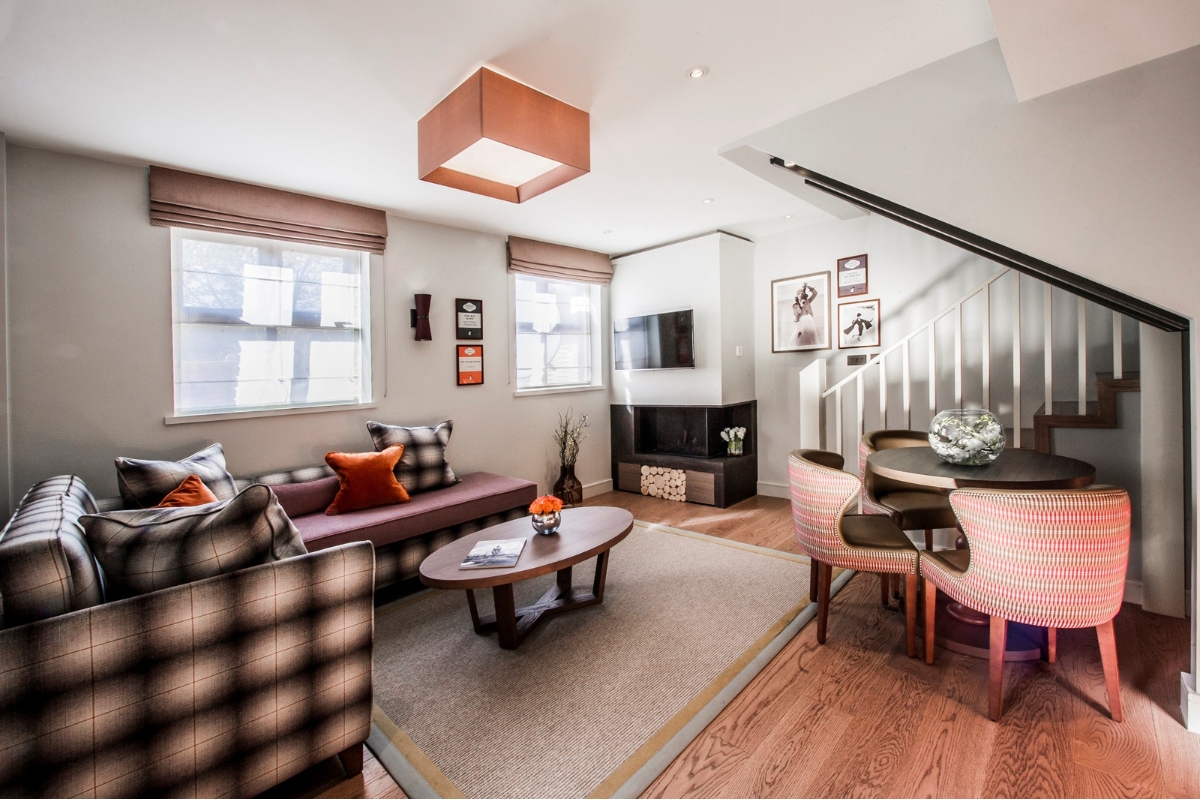 Modern multi-level hotel suite with plaid sofas, an oval coffee table, a small dining set, a fireplace corner, and a staircase leading upstairs.