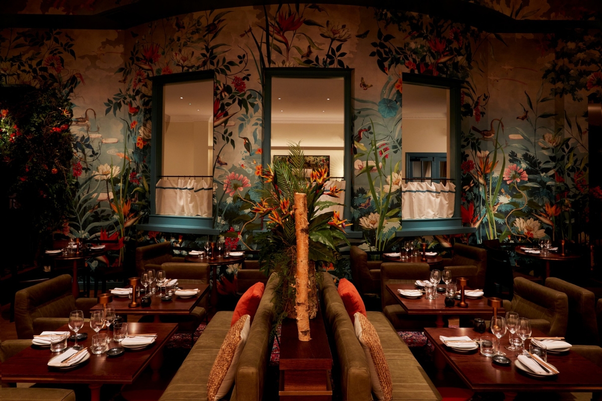 Dimly lit restaurant dining room with lush botanical wallpaper, set tables with stemware, and back-to-back green leather banquettes with orange throw pillows.