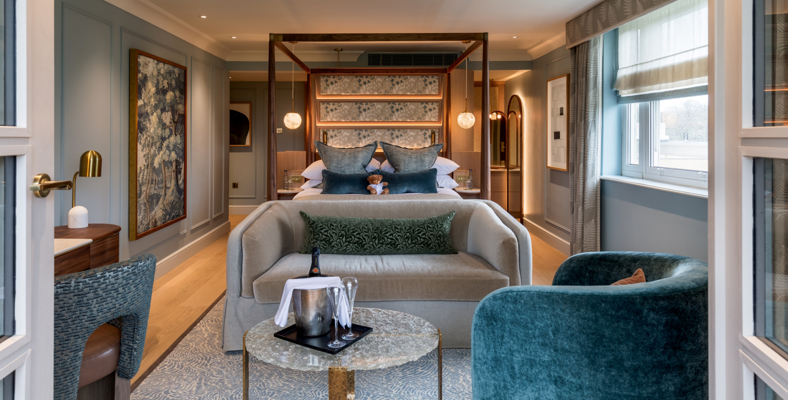 Elegant hotel room featuring a wooden canopy bed with warm glowing lights, a gray sofa at the foot of the bed, a glass coffee table with champagne, and soft blue walls.