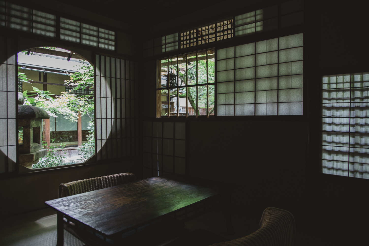 A moody, traditional Japanese tea room interior featuring a dark wooden table, wicker low seating, shoji sliding screens, and a large circular window framing a lush garden with a stone lantern.