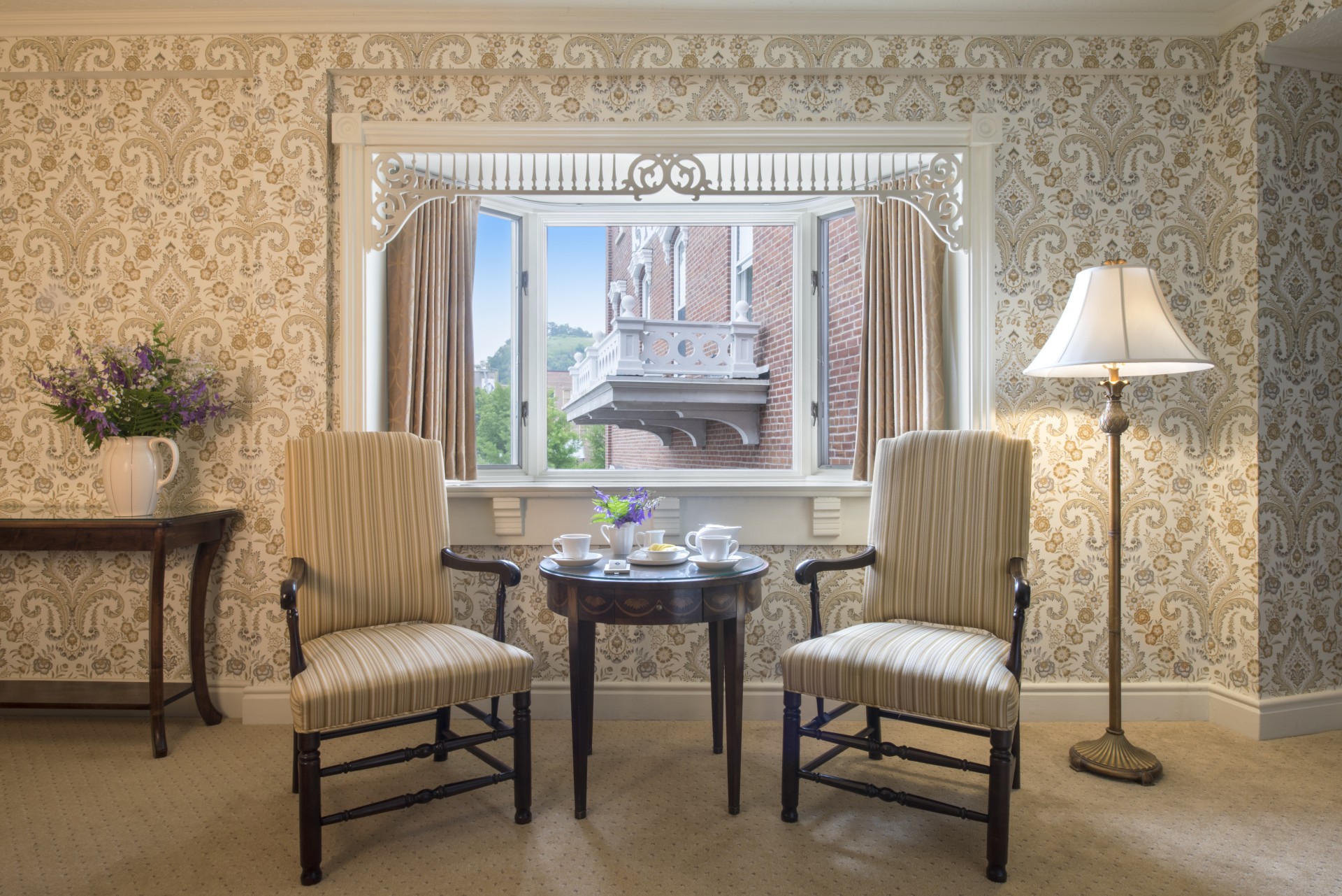 Cozy, vintage-style sitting alcove with detailed damask wallpaper, two striped armchairs framing a small wooden tea table, a standing lamp, and a bay window featuring ornate white fretwork trim looking out onto a brick building exterior.