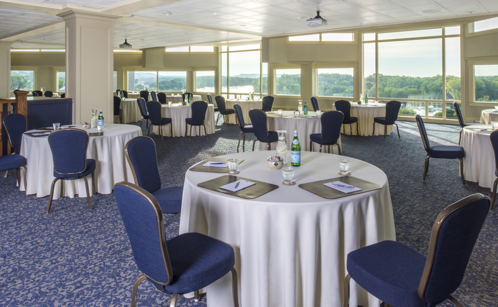 Spacious banquet or event hall set up with round tables, white tablecloths, and dark blue chairs, featuring expansive floor-to-ceiling windows with scenic views of a river and lush greenery.