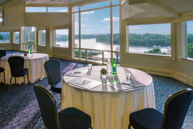 Bright conference room setup featuring round tables with white tablecloths and dark blue chairs, surrounded by large panoramic windows overlooking a sunlit river and wooded landscape.